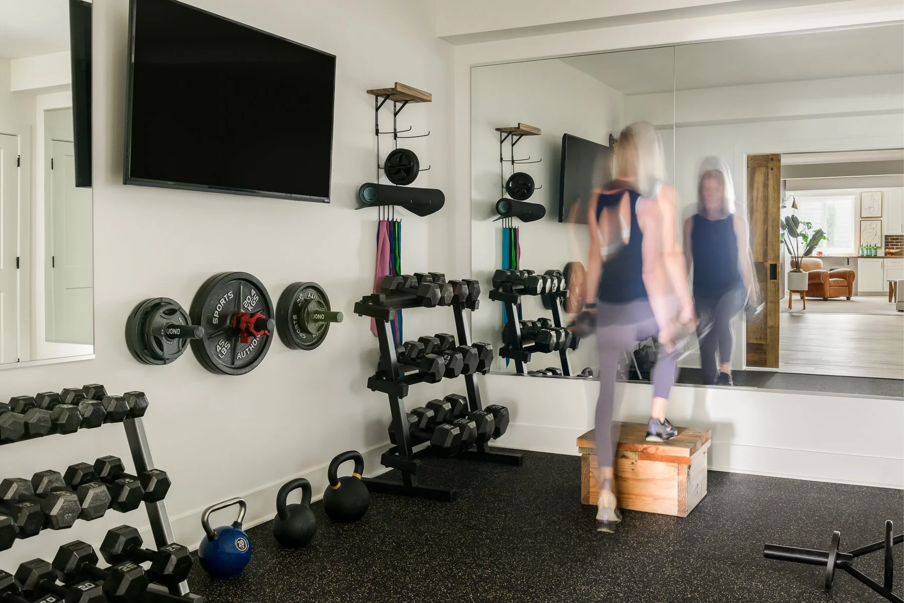 A woman with blonde hair in workout gear stands on a wooden box, lifting weights in a home gym with a large mirror, black dumbbells, kettlebells, and a TV on the wall.