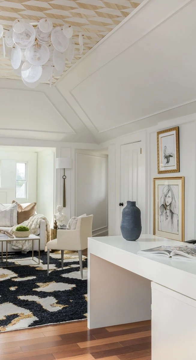 Bright living room with white walls, a black and white patterned rug, a beige sofa with pillows, a white armchair, a black vase on a white side table, framed artwork on the wall, a white desk with a blue vase and open magazine, and a chandelier hangi