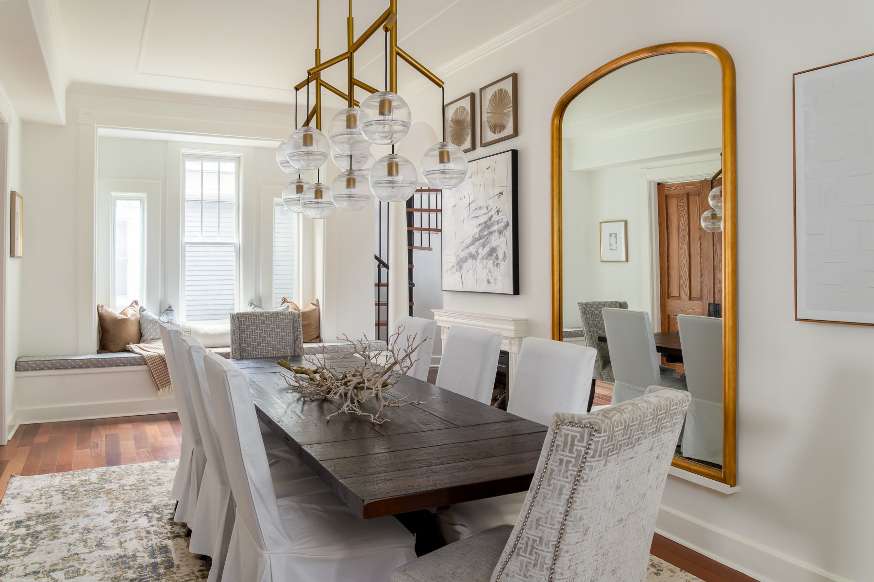 A dining room with a large wooden table, cream-colored upholstered chairs, and a decorative branch centerpiece. There is a built-in window seat with pillows near the windows and wall art. A gold-framed mirror hangs on the wall, reflecting part of the