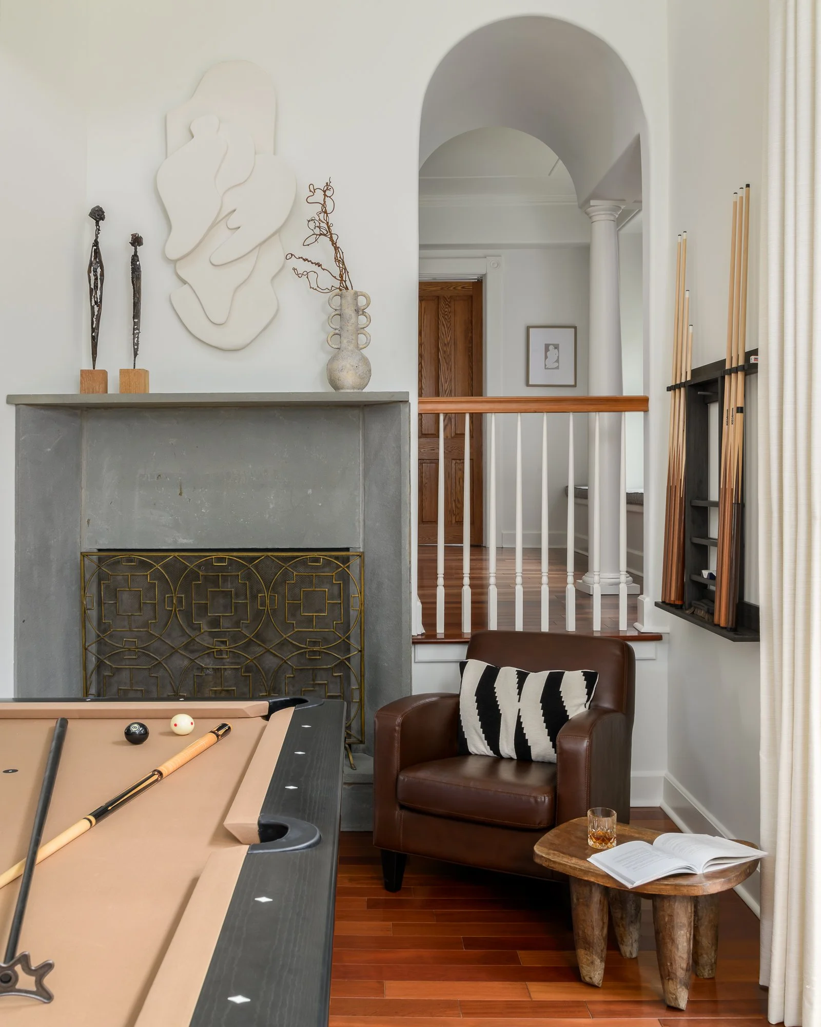 Interior view of a living space with a pool table, a brown armchair with a black and white pillow, a wooden side table with a glass of drink and an open book, a fireplace with a decorative screen, a staircase with painted white railings, a framed art