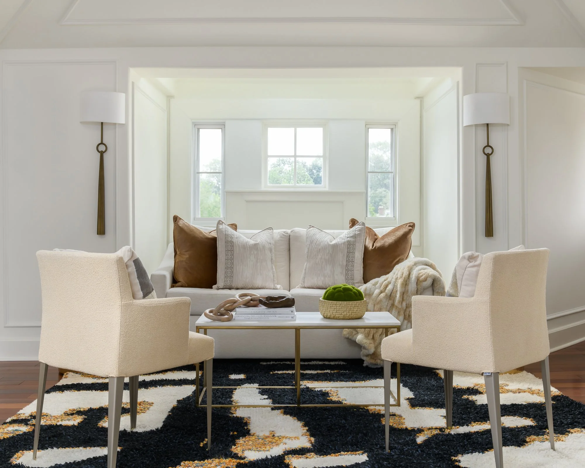 A modern living room with white walls, a large window letting in natural light, a white sofa with various pillows, an armchair, a coffee table with decorative items, and two wall-mounted lamps on each side.