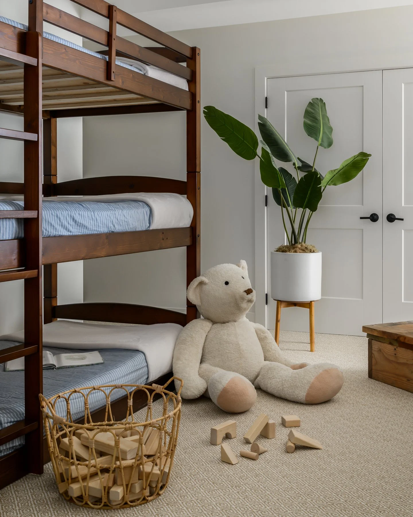 A bedroom with a wooden bunk bed, a large teddy bear, a potted plant with broad green leaves, a woven basket filled with wooden blocks, and scattered wooden blocks on a beige carpet.