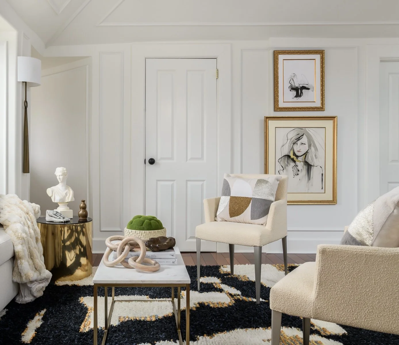 A cozy living room corner with white walls and a door. Decor includes artwork, a bust sculpture on a gold side table, a striking black and white rug, a modern beige armchair with a geometric pillow, another beige chair, a white coffee table with deco