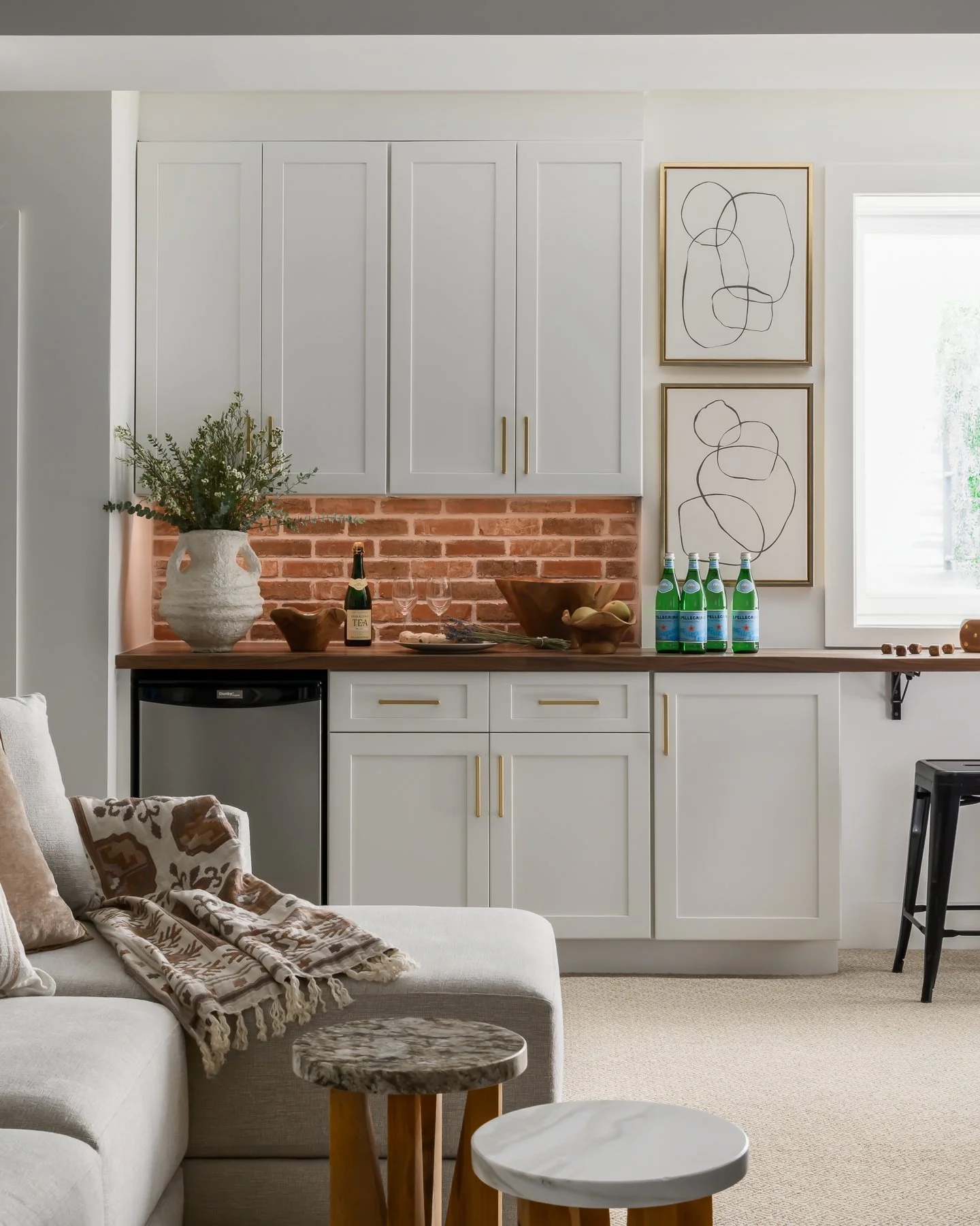 Living room with white sofa, decorative throw blanket, small round marble-top side tables, kitchen area with white cabinets, brick backsplash, framed abstract art, greenery, and bottled water