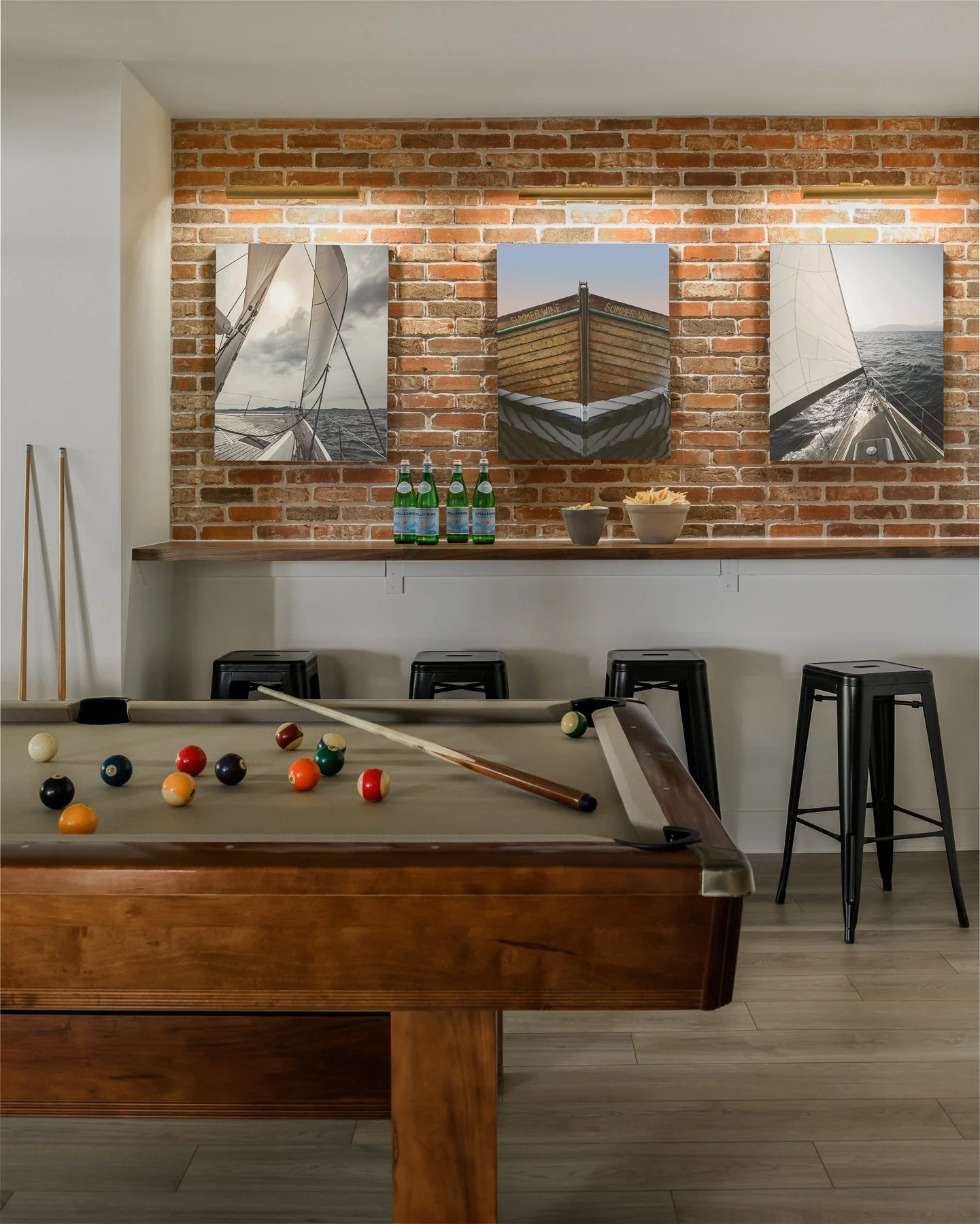 Interior of a room with a pool table in the foreground and a bar counter with barstools against a brick wall. Four bottles of sparkling water and two bowls of potato chips are on the counter. Three paintings of sailboats are hung on the brick wall, i