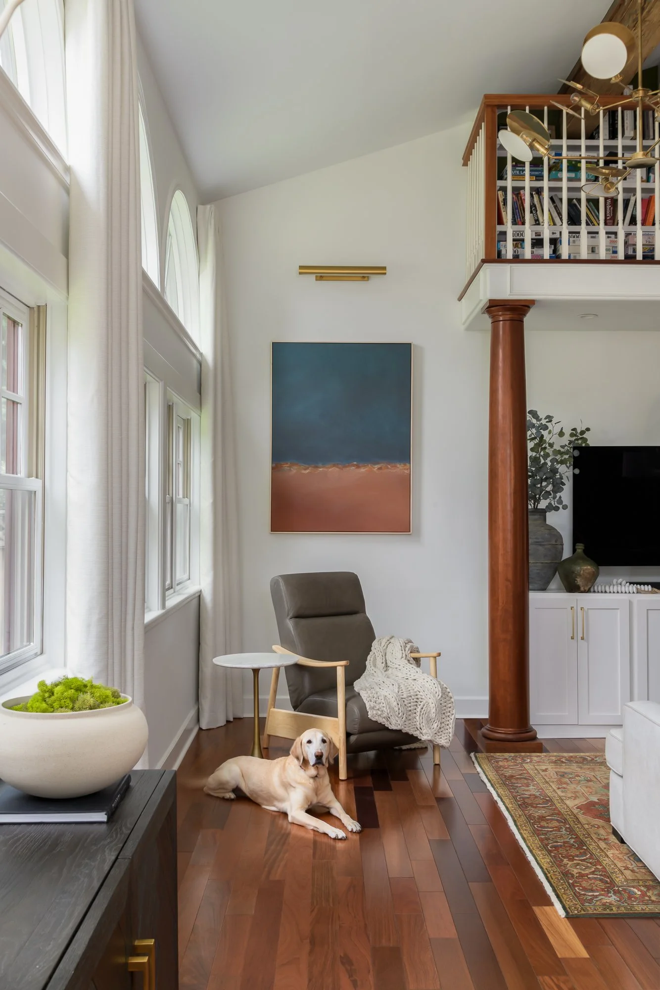 A cozy living room with a large window, white curtains, and hardwood floors. There is a gray armchair with a knitted blanket and a small side table, a Labrador retriever lying on the floor, and a wooden console table with a decorative plant. A large 