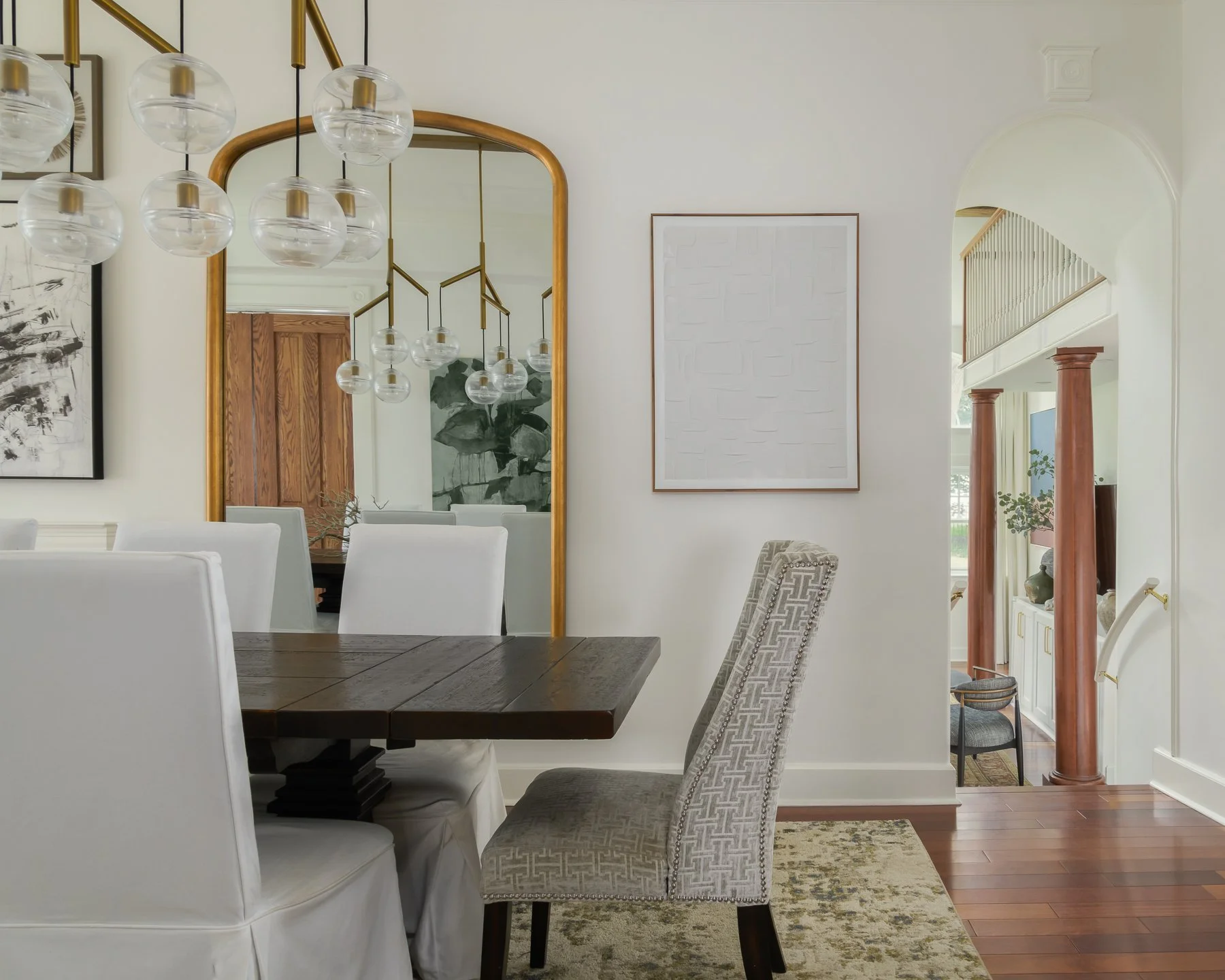 A dining room with a dark wood table, white upholstered chairs, and a patterned chair in the foreground. A large gold-framed mirror and modern chandelier with glass globes are on the wall. There are framed art pieces, one with black and white abstrac