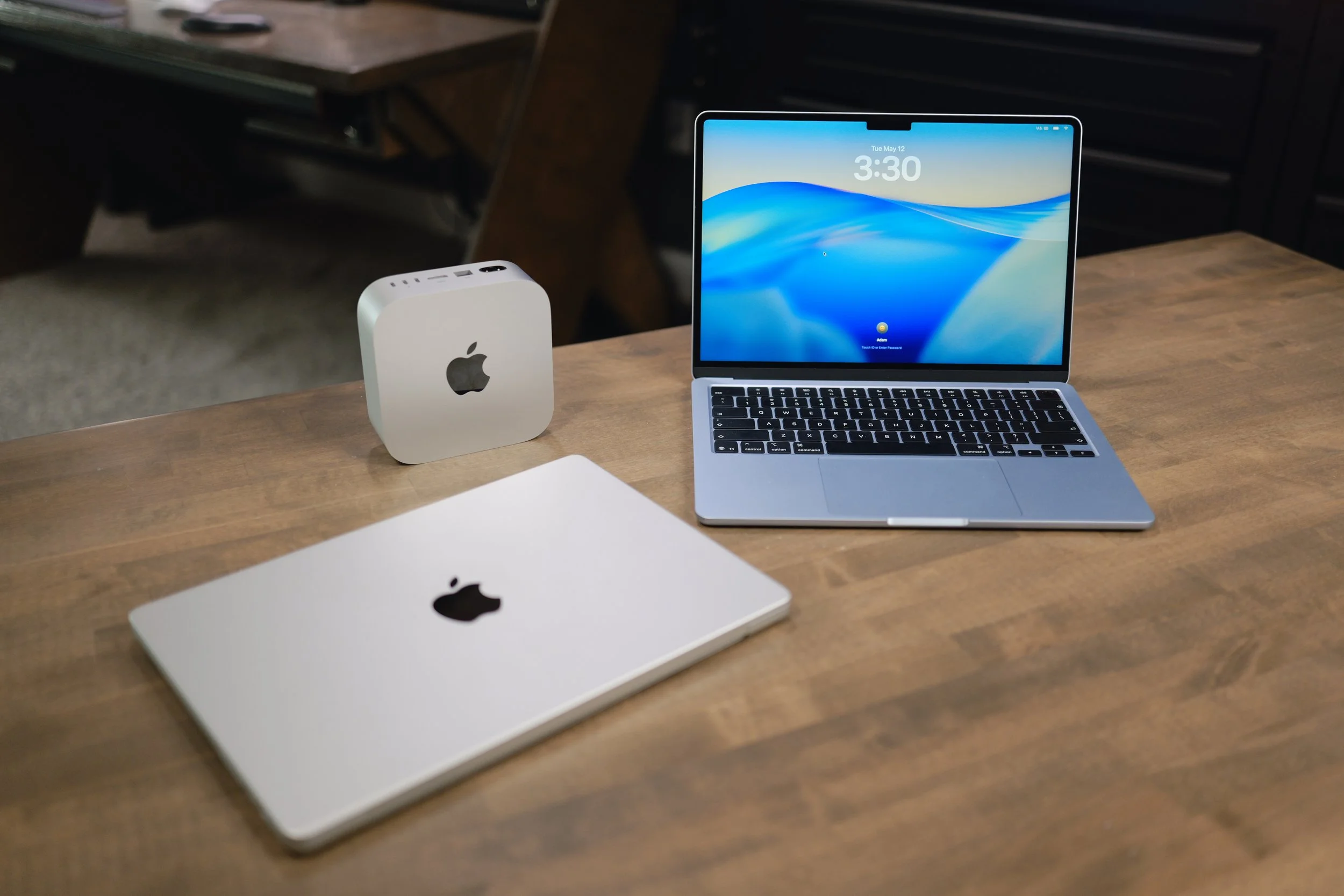 An open laptop with a blue screen displaying the date and time, alongside a white Apple Mac mini and a closed white MacBook on a wooden table, with a dark office background.