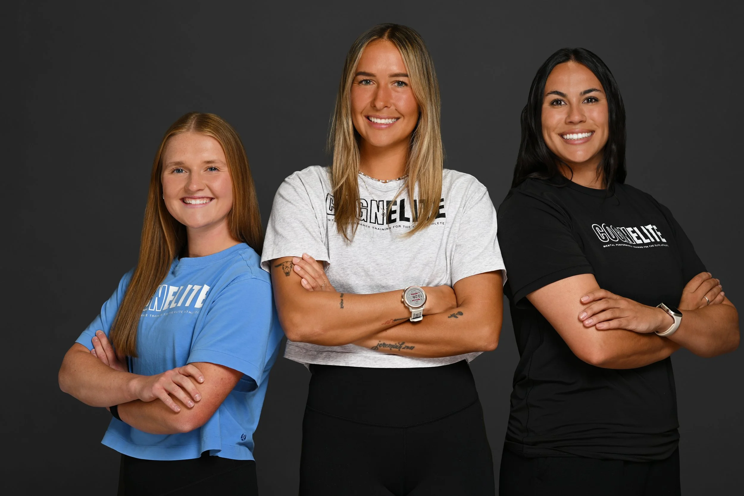 Three women standing side by side with arms crossed, smiling, against a dark background. They are wearing casual athletic t-shirts, with logos related to G.O.O.ELITE, a mental performance training organization.