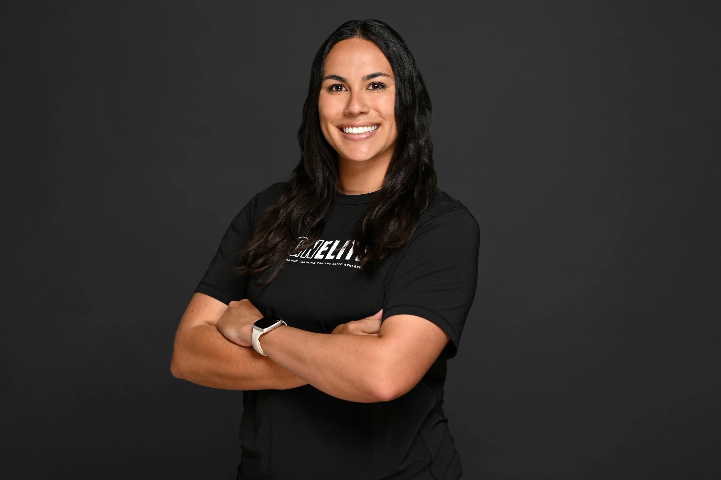 A woman with long dark hair smiling with arms crossed, wearing a black T-shirt and a smartwatch, standing against a dark gray background.