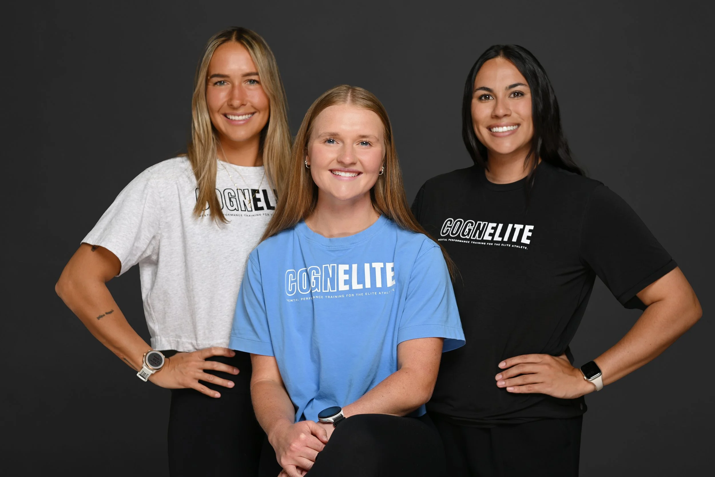 Three women standing against a dark background, smiling at the camera. They are wearing casual t-shirts with the word 'COGNELITE' printed on them, indicating they are part of an athletic or training team.