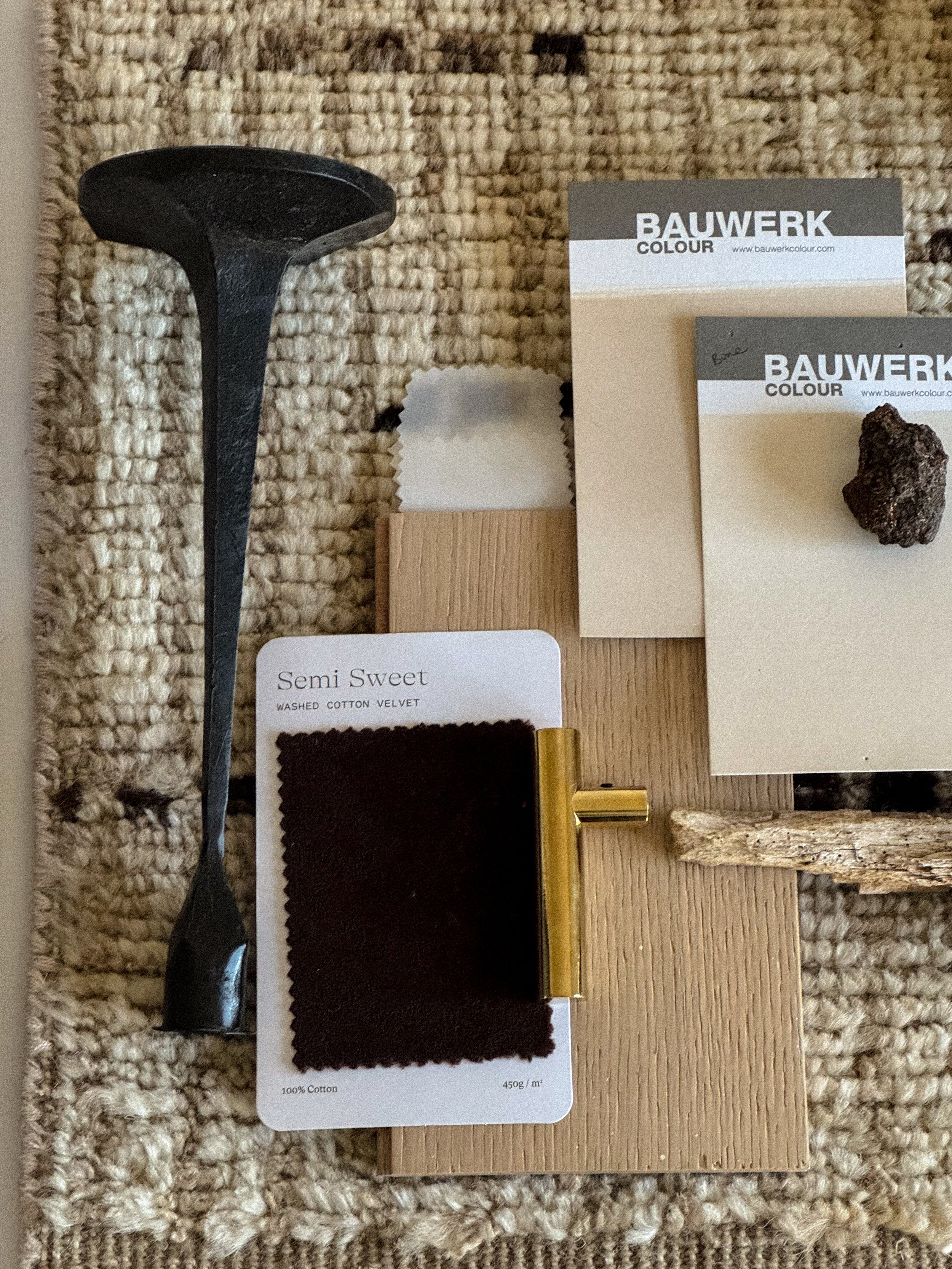 Color palette samples with black fabric swatch labeled "Semi Sweet," a small piece of dark fabric, wooden pieces, and a small rock, arranged on a beige textured surface.
