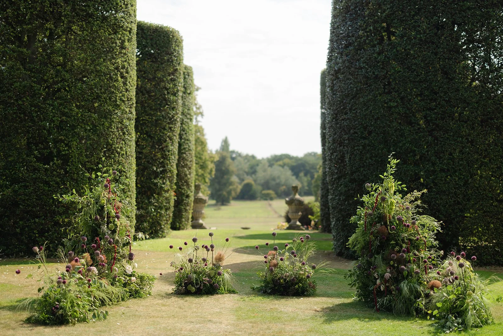 A garden pathway framed by tall, neatly trimmed hedges on both sides, with flower arrangements at the entrance, leading to decorative urns in the distance, under a bright sky.