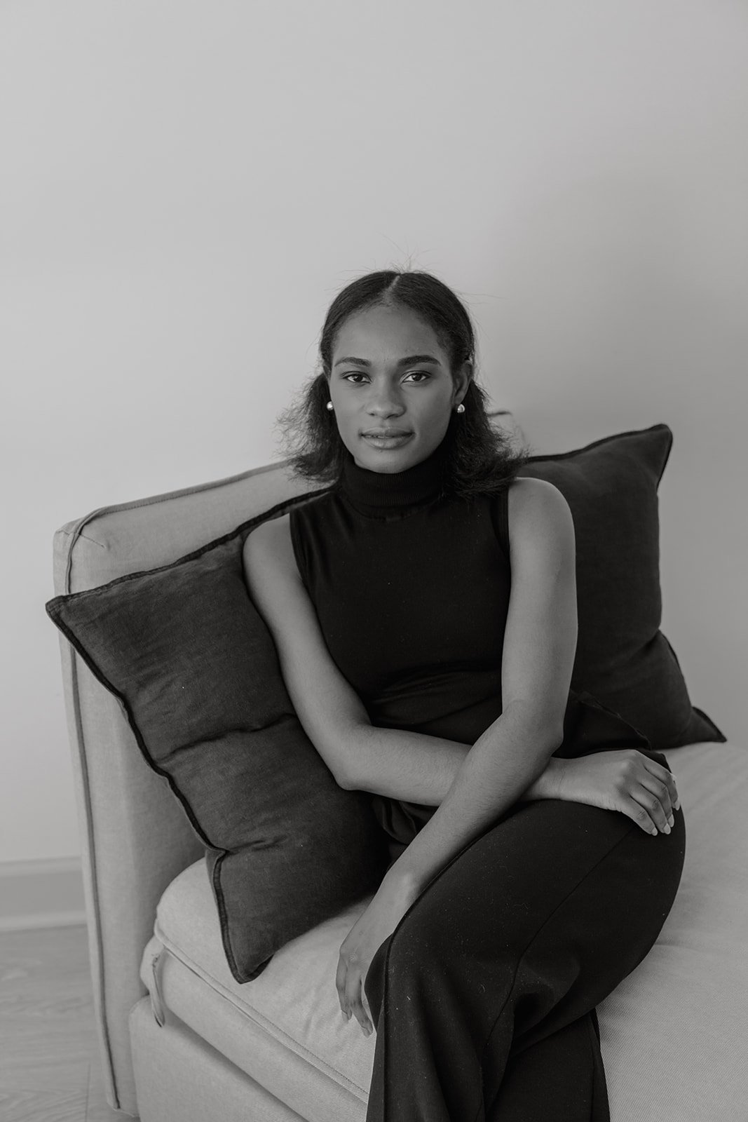 A woman sitting on a couch with pillows behind her, wearing a sleeveless black turtleneck top and earrings, looking at the camera in a black and white photo.