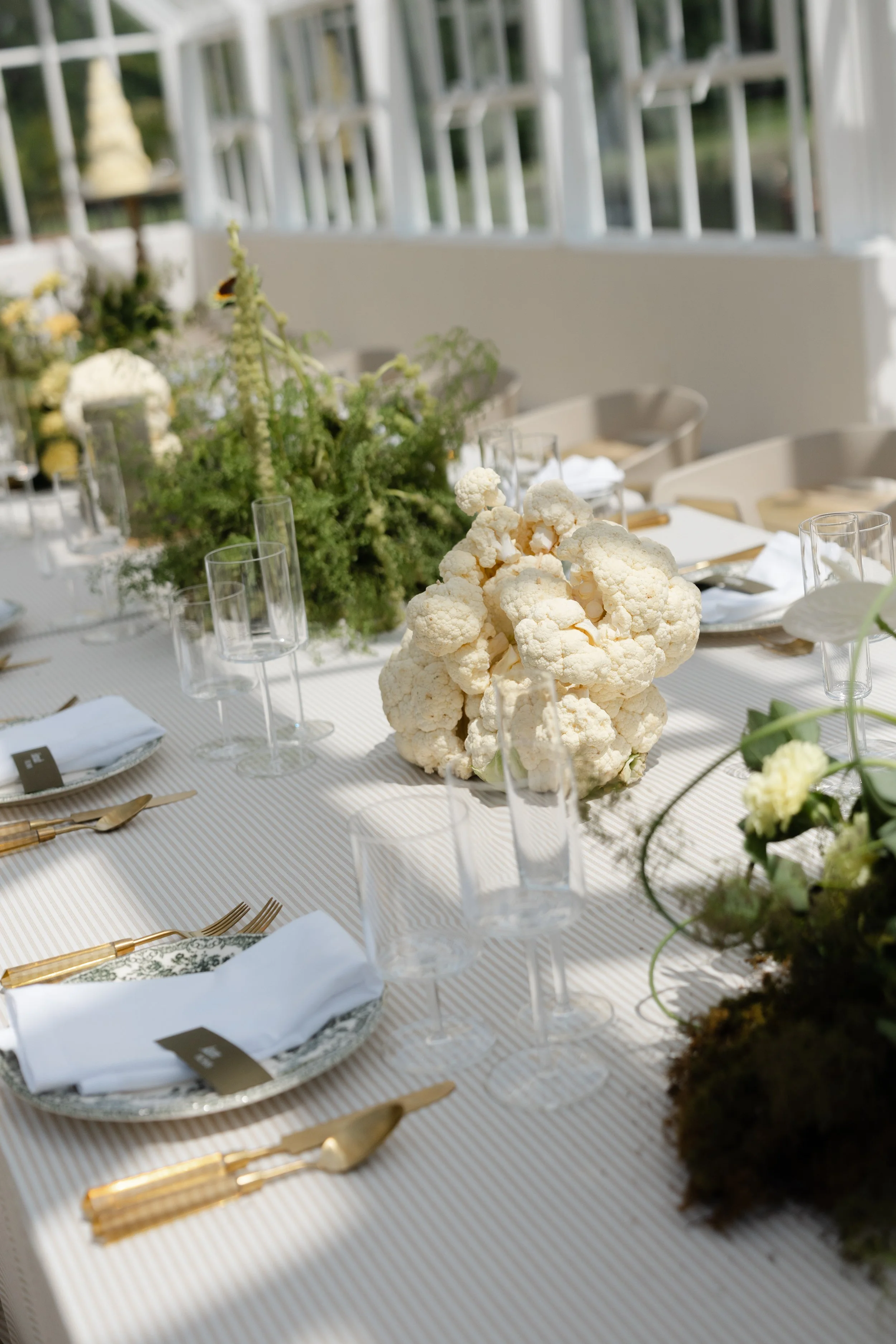 A formal dining table decorated with a large cauliflower centerpiece, greenery, and white and gold tableware in an outdoor setting with natural light.
