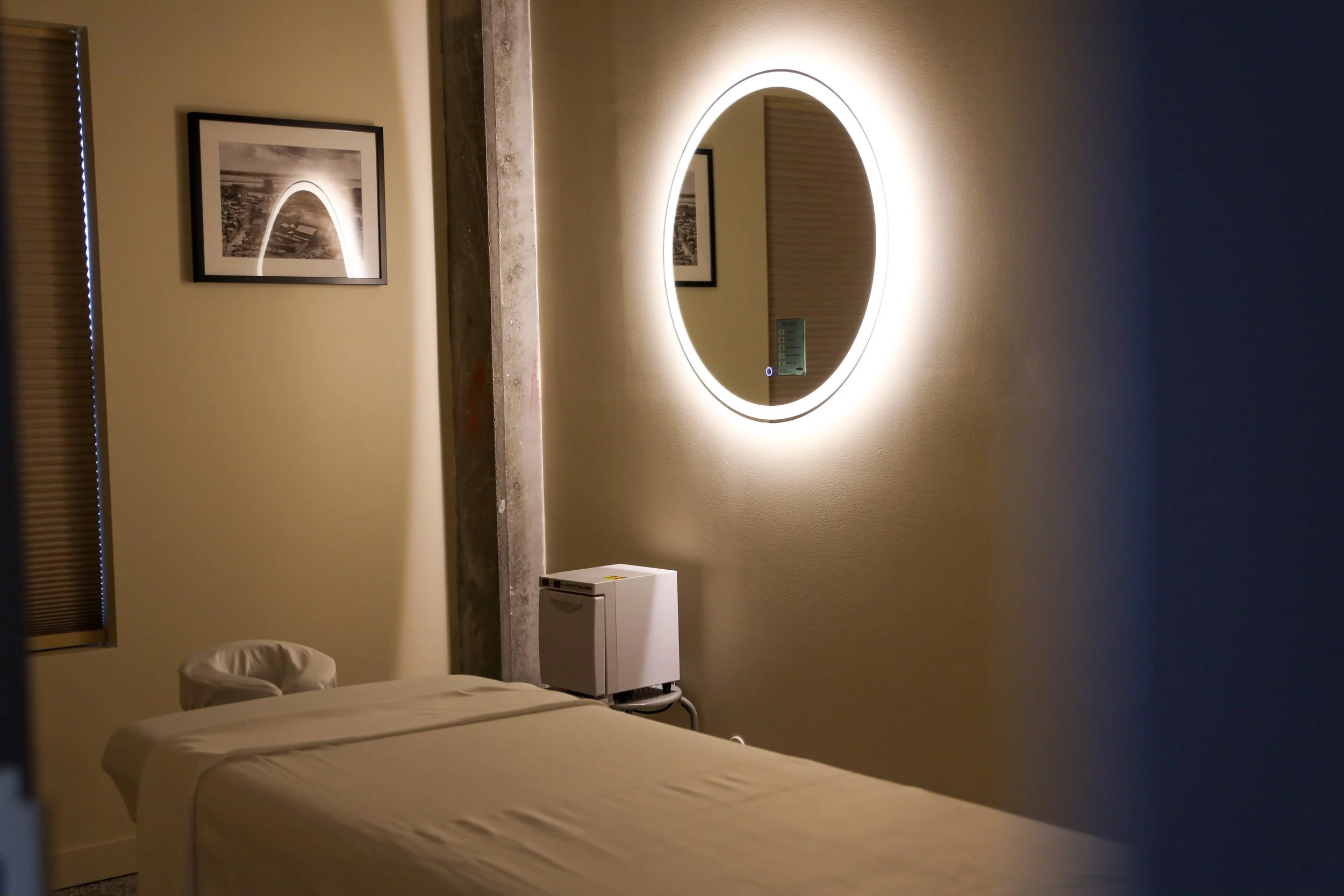 Une chambre avec un lit, un mur avec miroir rond éclairé, un tableau noir et blanc, une fenêtre avec volets, et un distributeur d'eau ou un appareil électroménager.