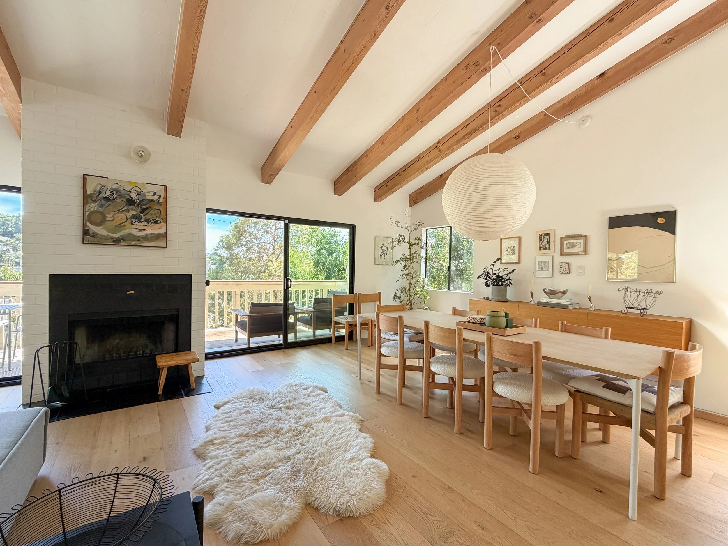 Light-filled Northeast Los Angeles Airbnb dining room with exposed wood beams, white oak floors, fireplace, large windows and warm wood furnishings.