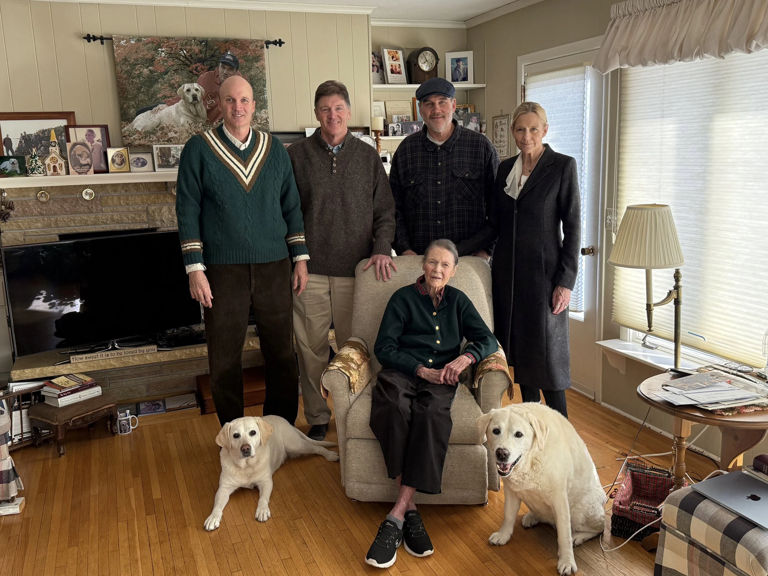 A family of six people, including an elderly woman sitting in a chair, four standing adults, and two Labrador retrievers, in a cozy living room with framed photographs and artwork on the walls.