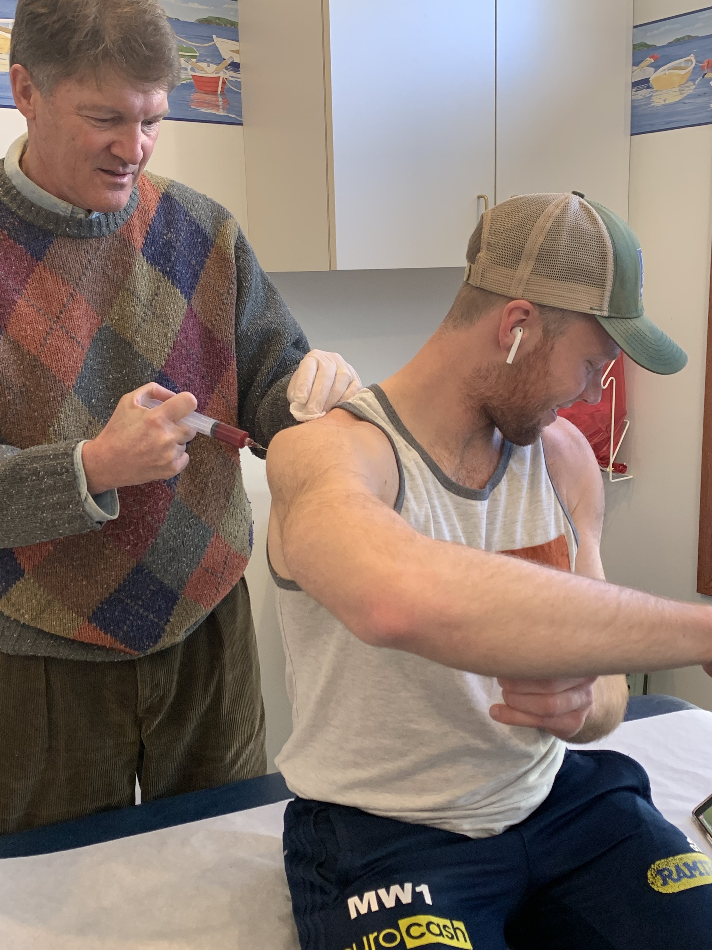 A man in a sleeveless shirt receiving an injection in his upper arm from another man in a sweater. The seated man is wearing wireless earbuds and a baseball cap, with a tattoo on his right arm. The scene appears to be in a medical or clinical setting.