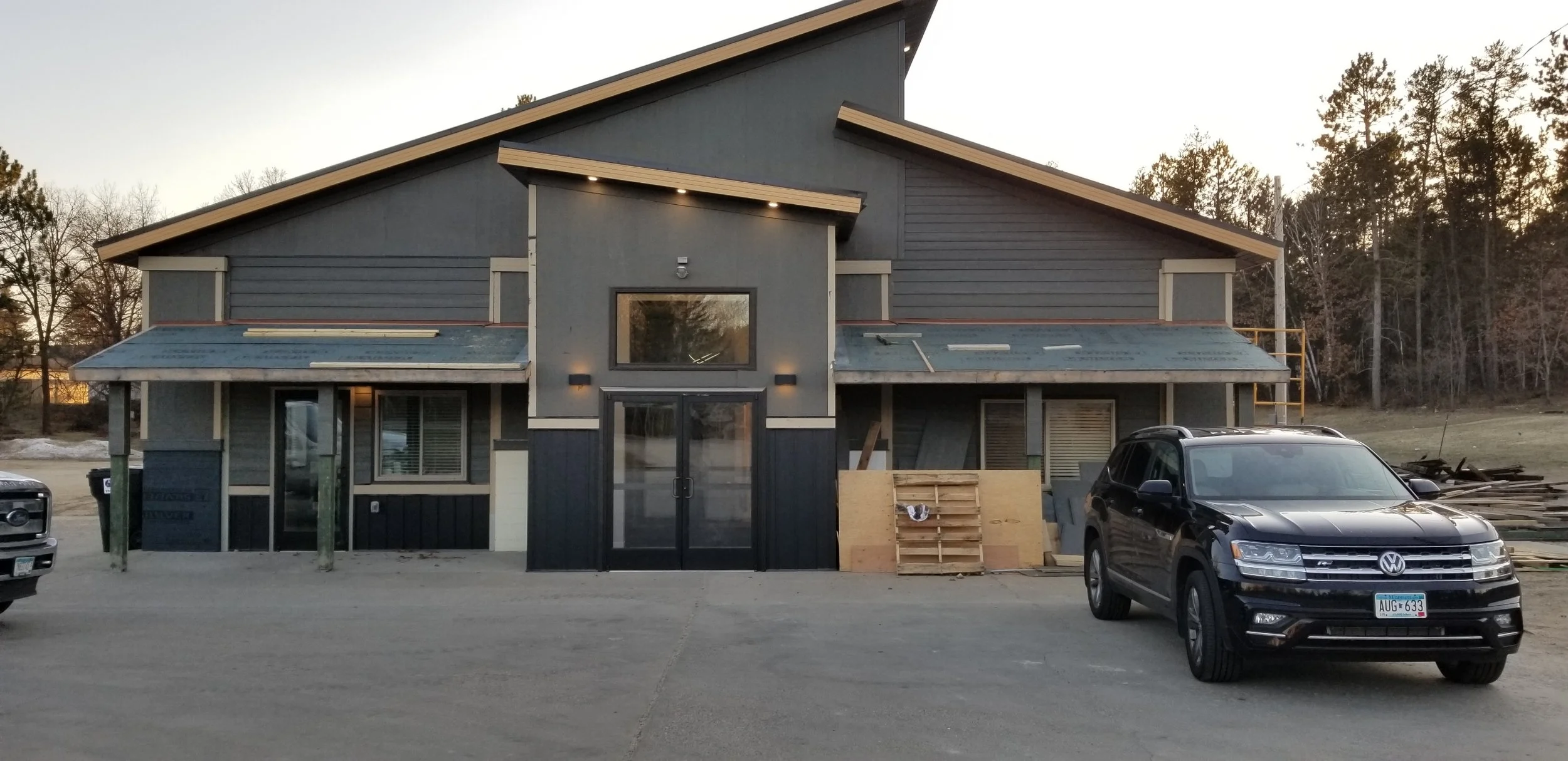 Under construction modern building with gray and beige exterior, large glass doors, and parked black Volkswagen SUV in front, surrounded by construction materials and an open area with trees in the background.