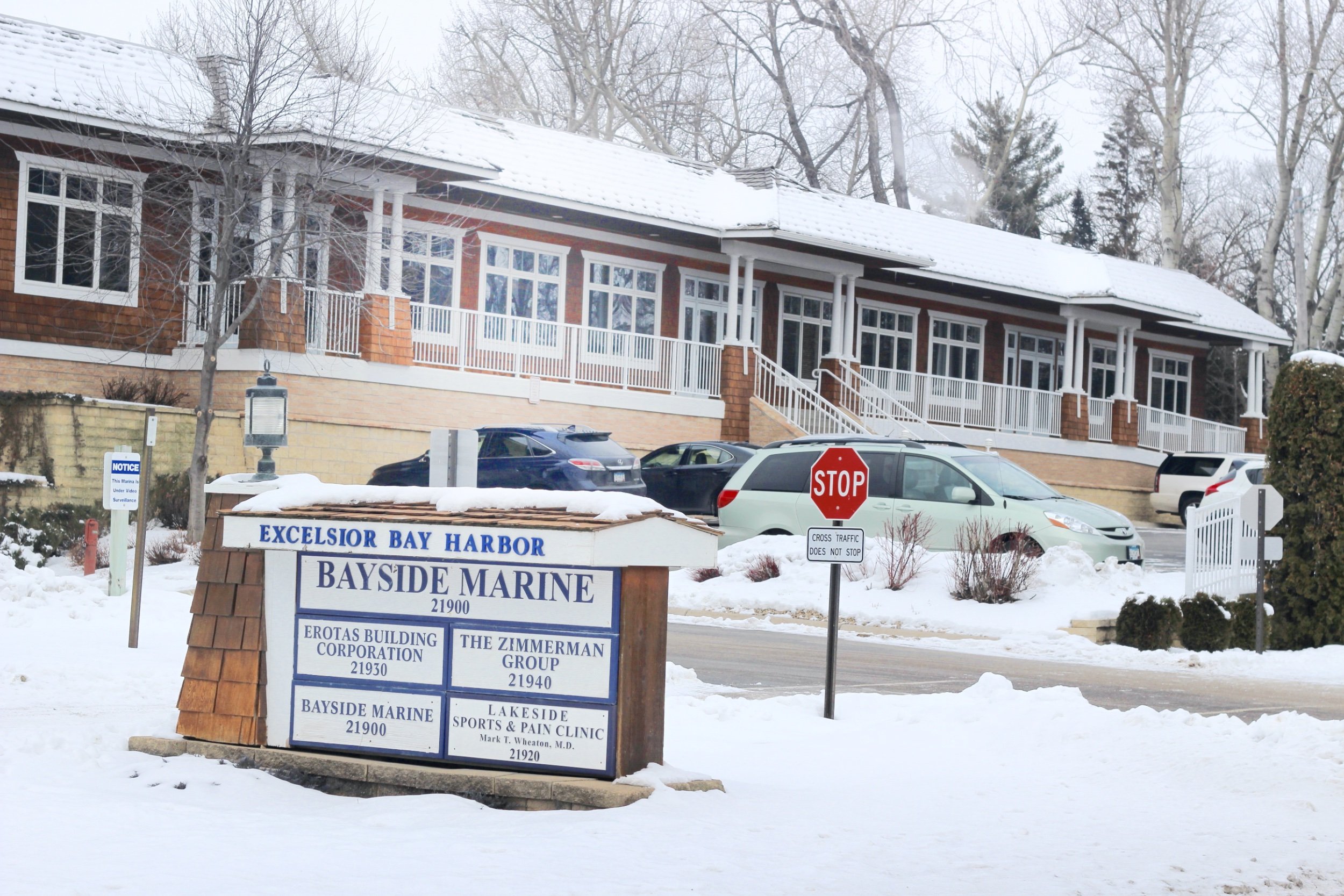 Snow-covered entrance to a building with a sign for Bayside Marine, Erotas Building Corporation, and Lakeside Sports & Pain Clinic, with parked cars and a stop sign in front.