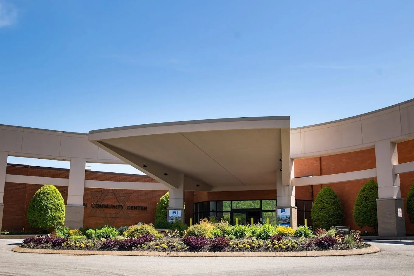 Front view of a community center with a landscaped roundabout in front, featuring green bushes and flowering plants, and a blue sky overhead.