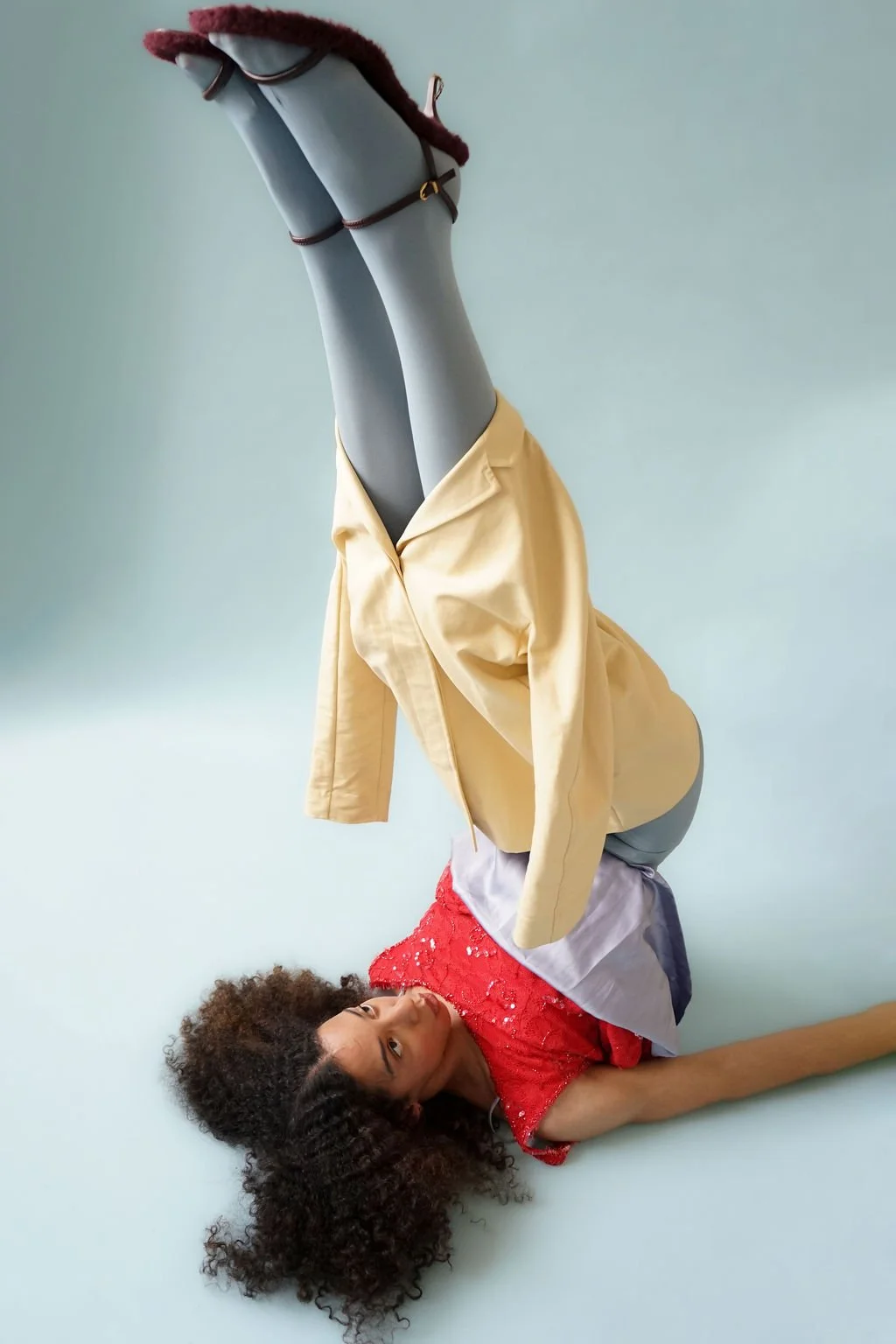 A woman with curly hair lying on the floor, looking at the camera, with her legs raised and resting on a mannequin wearing a beige jacket, gray pants, and a red textured top. The background is plain.