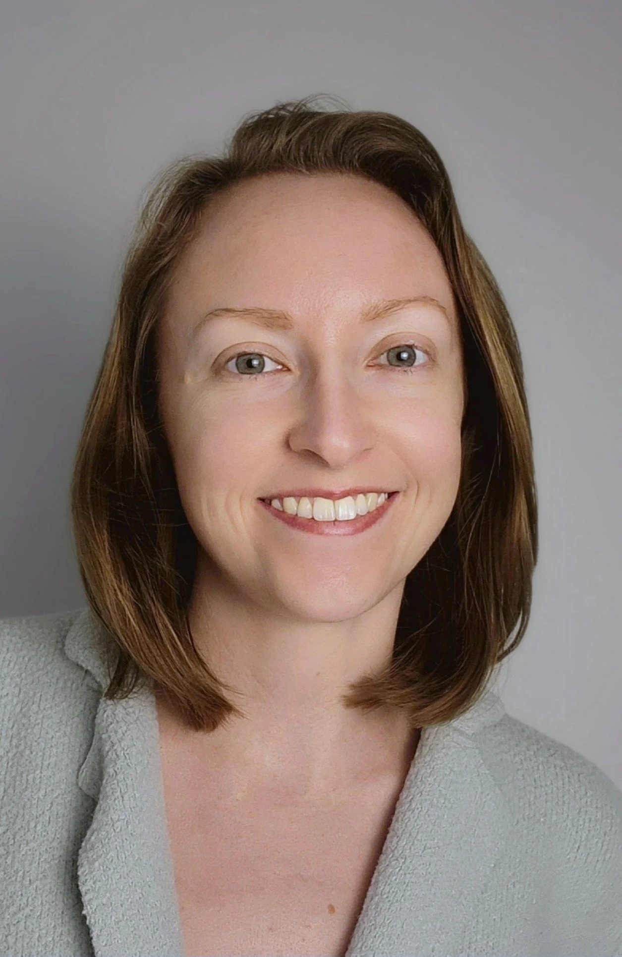 Close-up portrait of a smiling woman with shoulder-length brown hair, wearing a light gray coat, against a plain gray background.