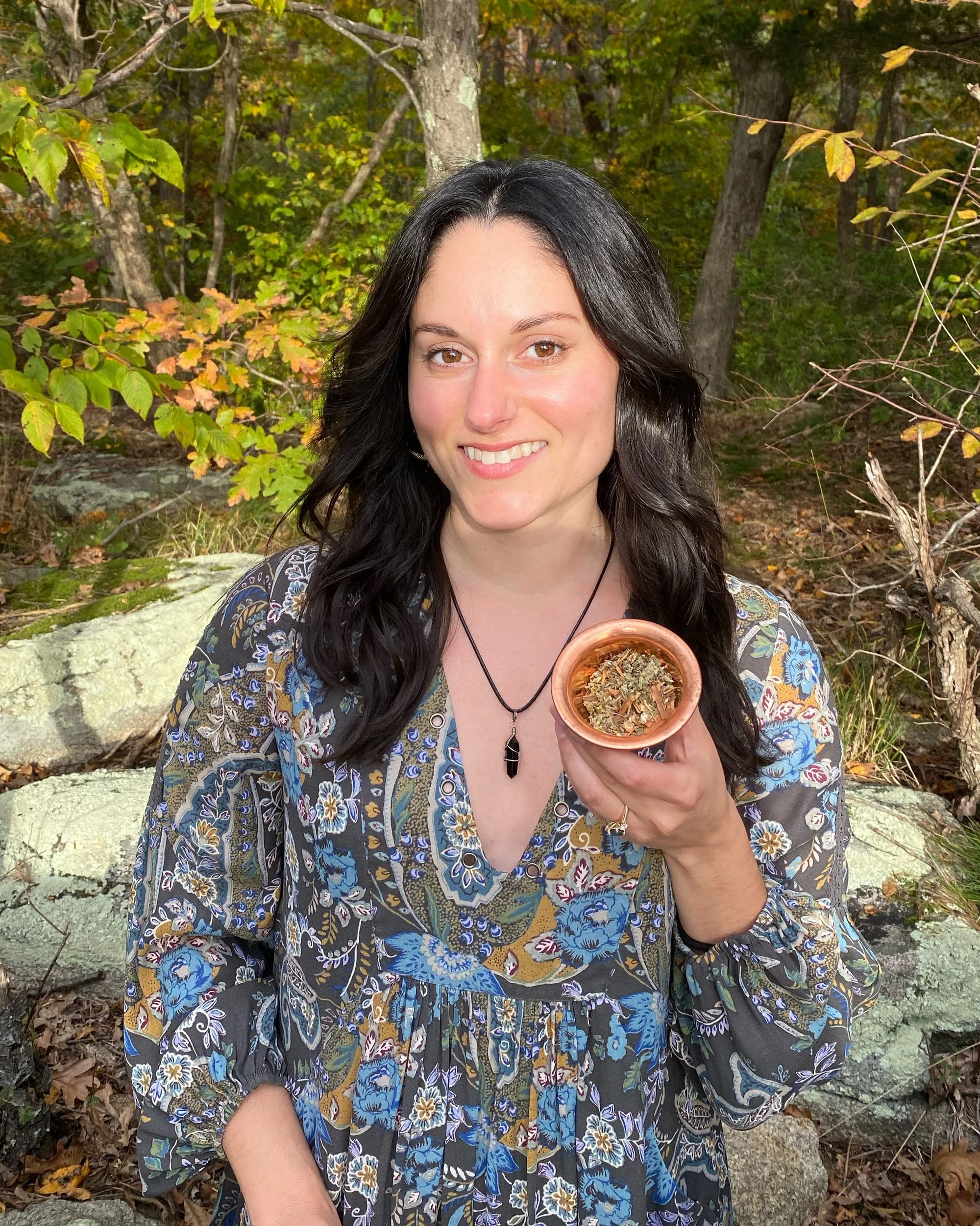 A woman with long dark hair smiling outdoors in a wooded area, holding a small bowl filled with dried herbs or plants.