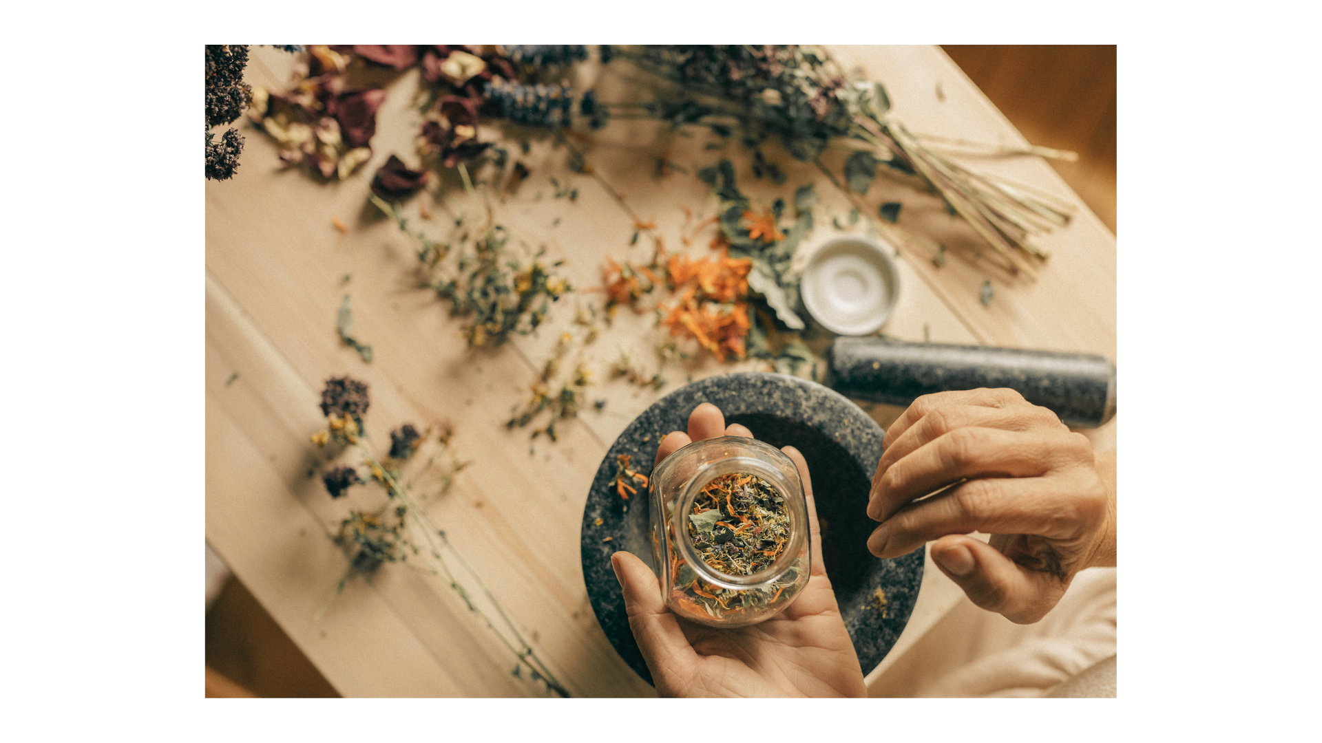hands holding a glass jar full of loose herbs with a mortar and pestle on the table and more dried herbs and flowers.