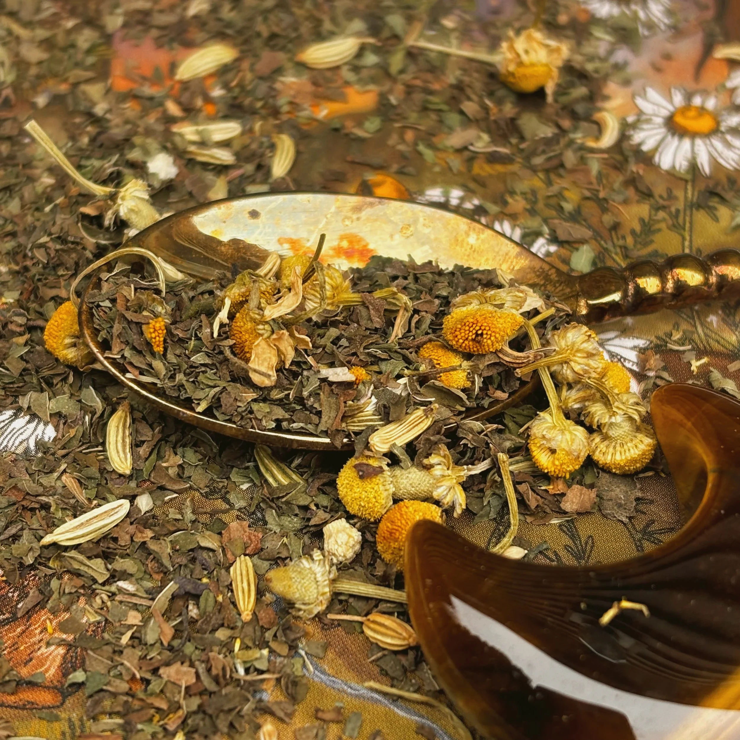 loose leaf herbs of peppermint, fennel, ginger and chamomile on a gold spoon with a tigers eye crescent moon shaped crystal