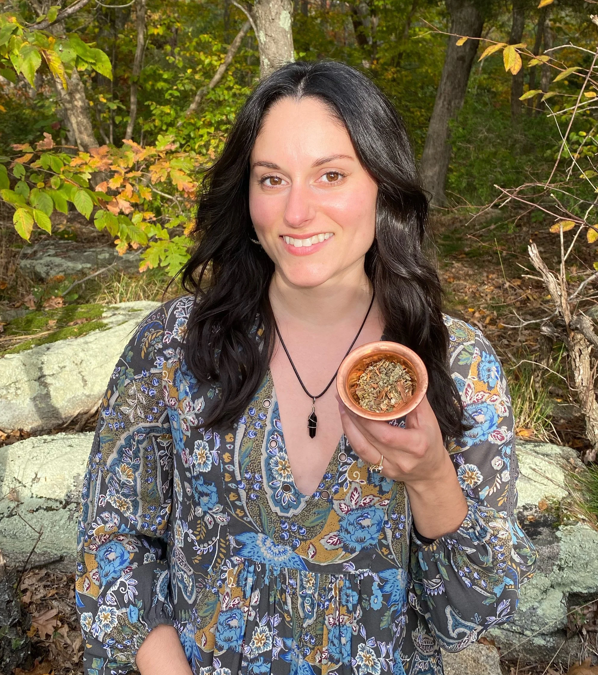 a woman with long heair smiling and holding a bowl of loose leaf herbal tea in the woods with trees and rocks on the background