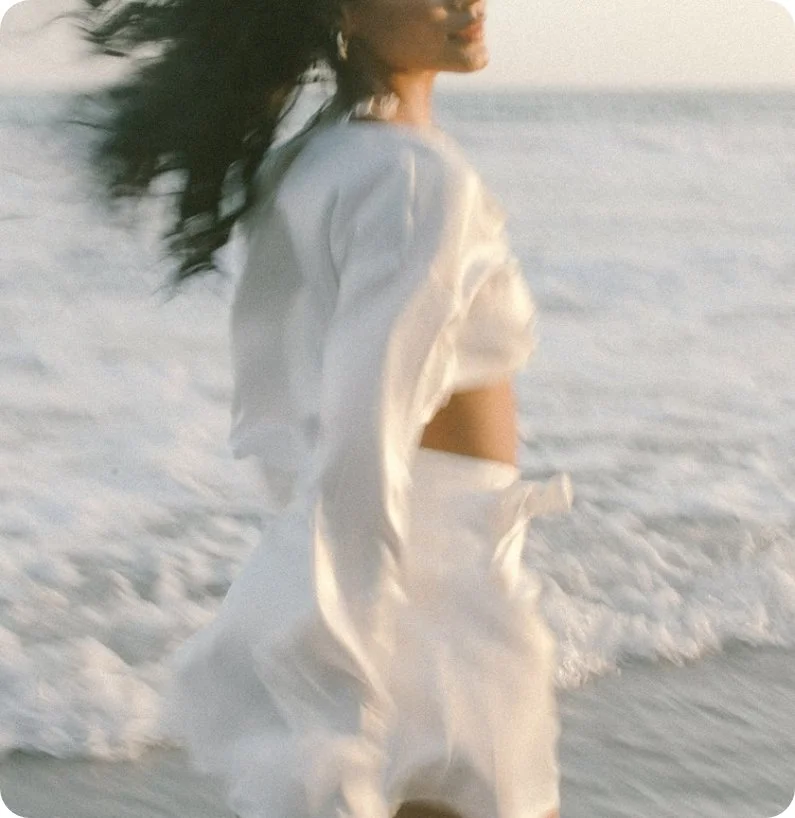 A woman standing in the ocean, wearing a white outfit, with wind-blown hair, facing sideways.