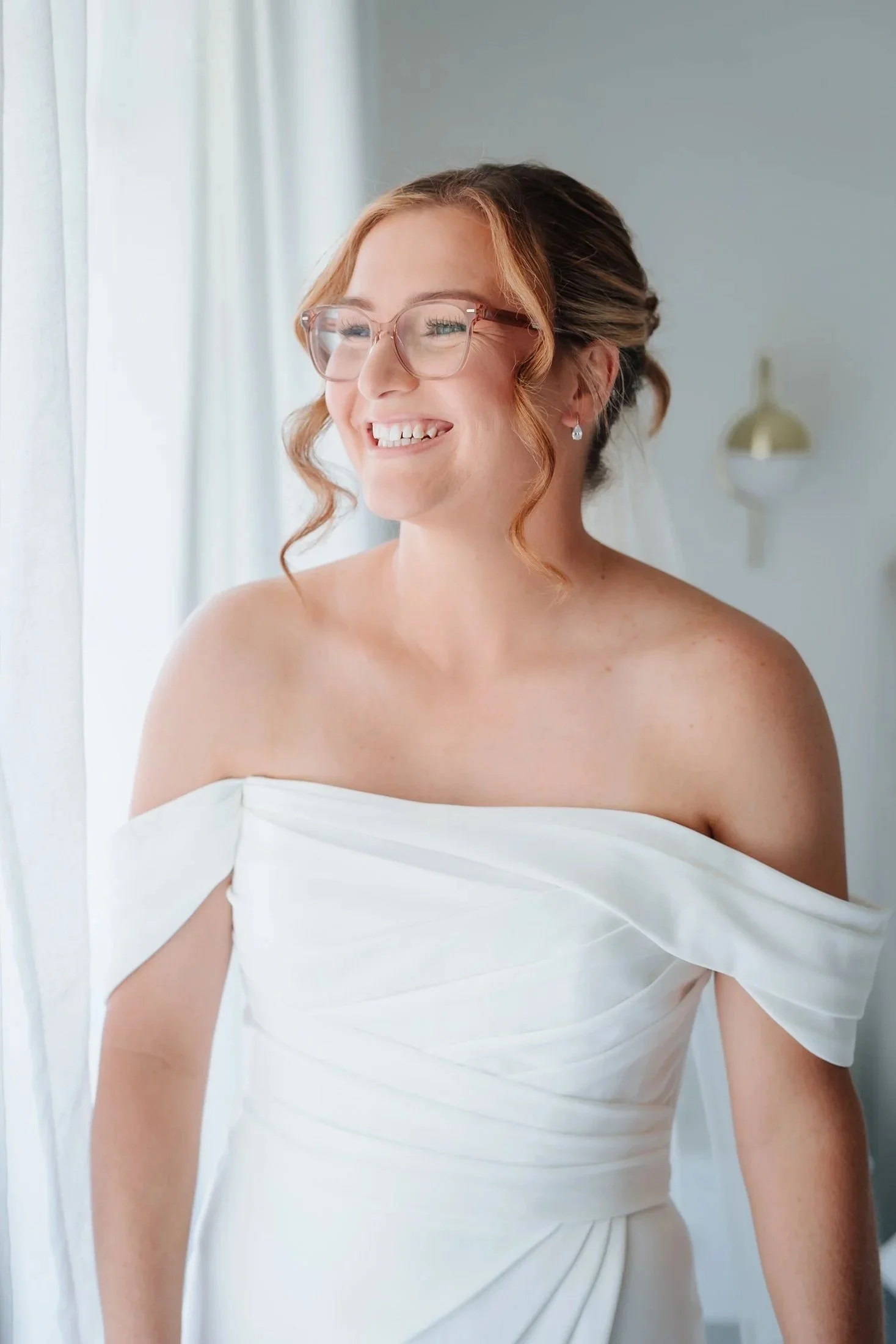 A smiling bride with glasses in a white off-the-shoulder wedding dress, standing near a window with white curtains.