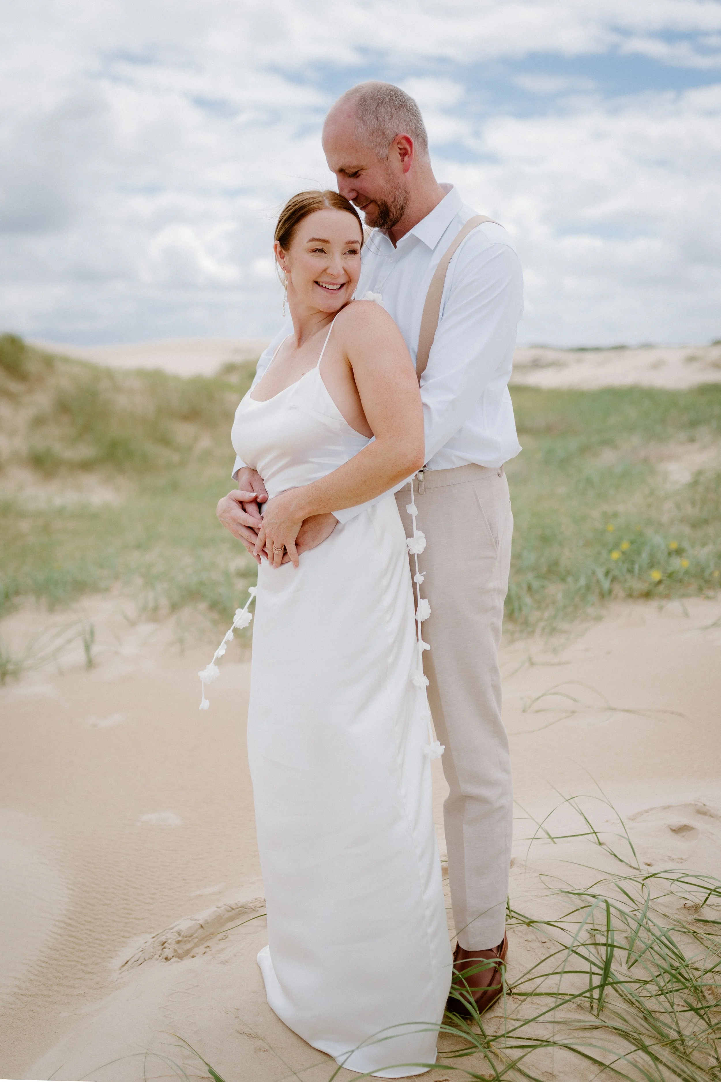 A couple on a sandy beach, dressed in white wedding attire, embracing happily under a cloudy sky.