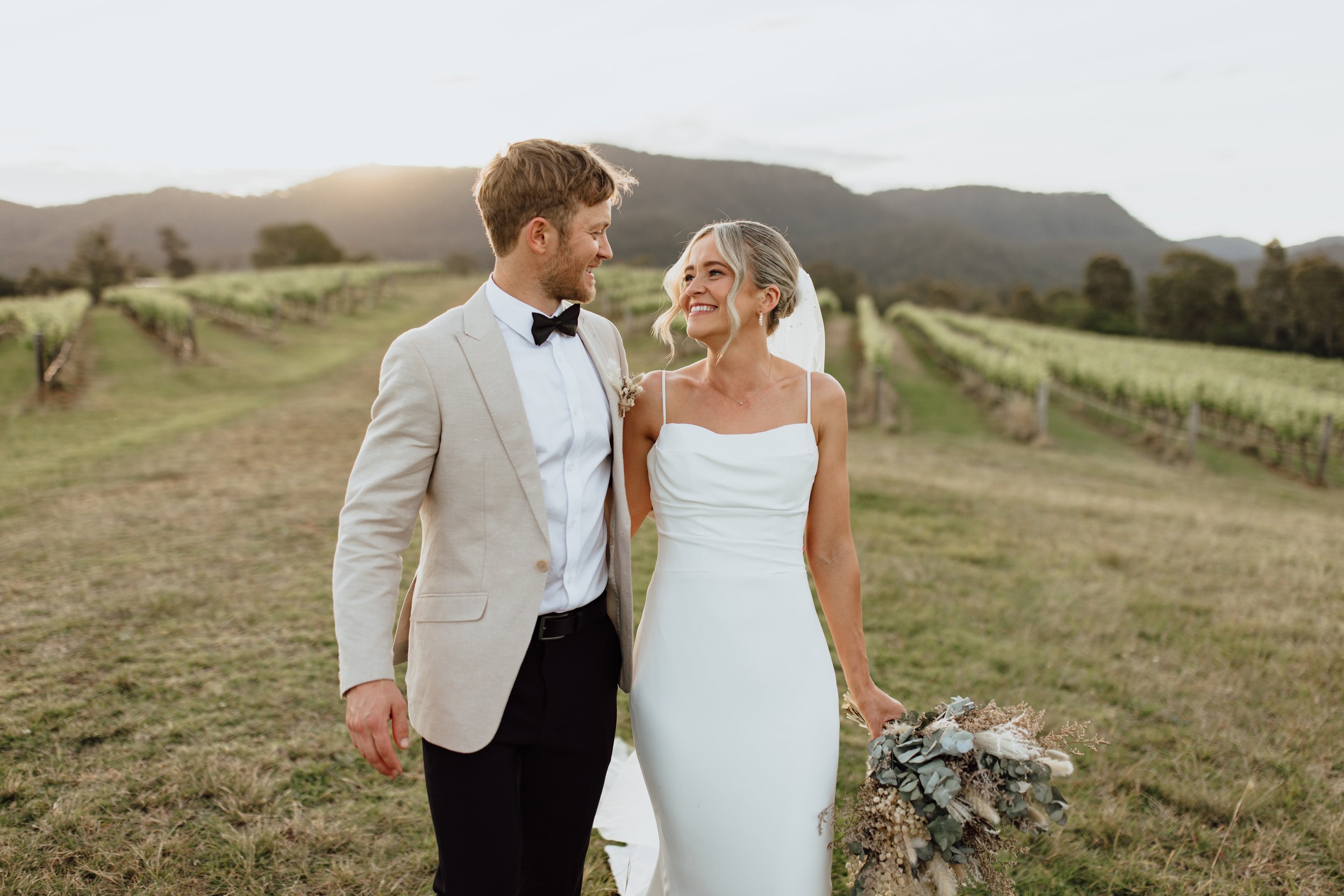A bride and groom smiling at each other on a vineyard with mountains in the background during sunset.