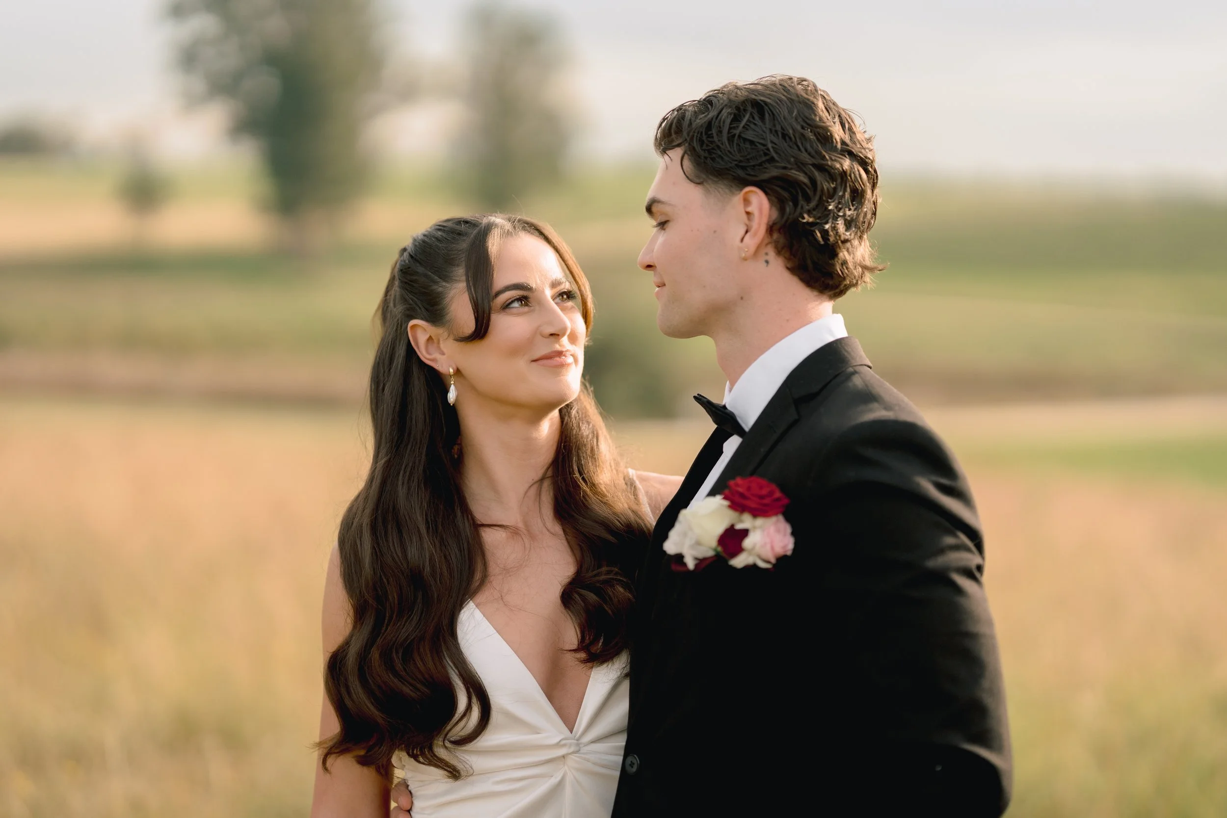 A couple dressed in wedding attire standing outdoors in a field, gazing at each other.