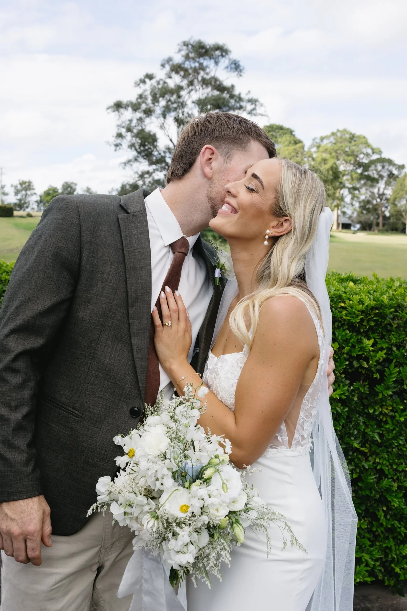A bride and groom sharing a kiss outdoors, with greenery and trees in the background. The bride holds a bouquet of white flowers.