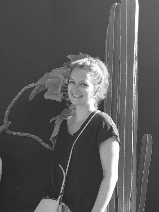 Jeune femme souriante avec un sac, debout devant un cactus et une plante artificielle, en noir et blanc.