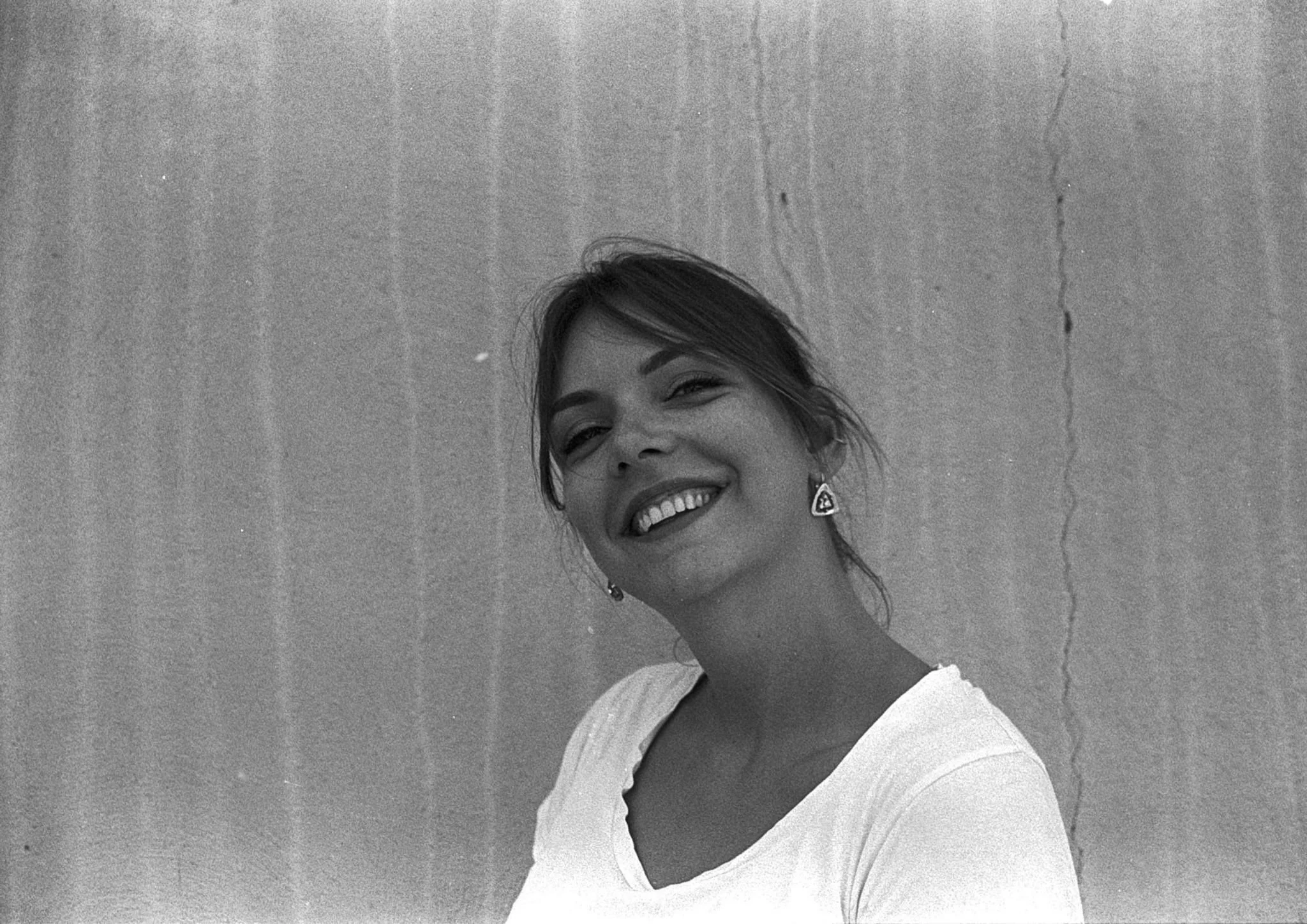 Portrait en noir et blanc d'une jeune femme souriante portant des boucles d'oreilles triangulaires, devant un mur en béton.