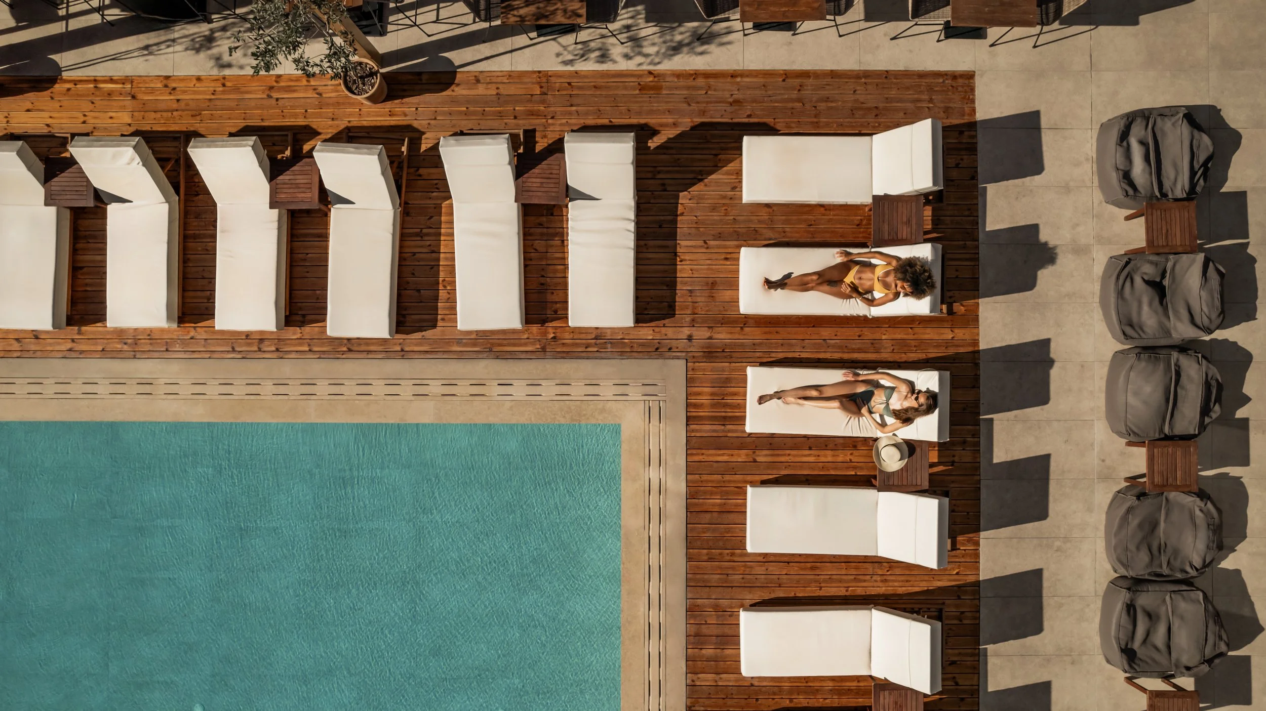 Two women sunbathing on lounge chairs beside a swimming pool, with a wooden deck and beige tiled area. One woman is lying on a white lounge chair wearing a yellow swimsuit, while the other woman is reclining on a similar lounge chair wearing a green bikini. There are some small tables, large gray bags, and closed umbrellas nearby.
