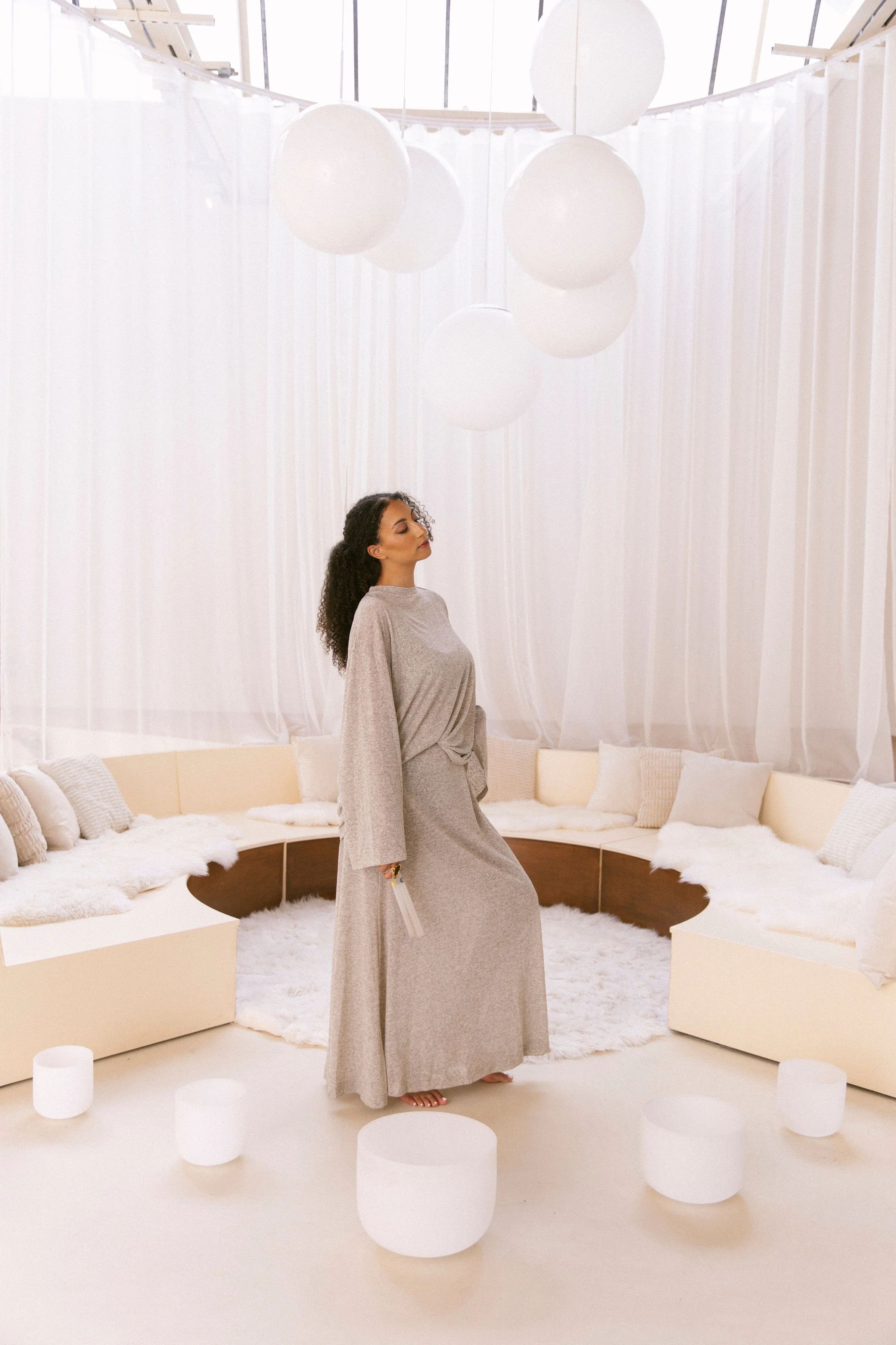A woman standing in a modern, minimalist, circular sitting area with white sofas, pillows, and soft lighting from hanging spherical lamps.