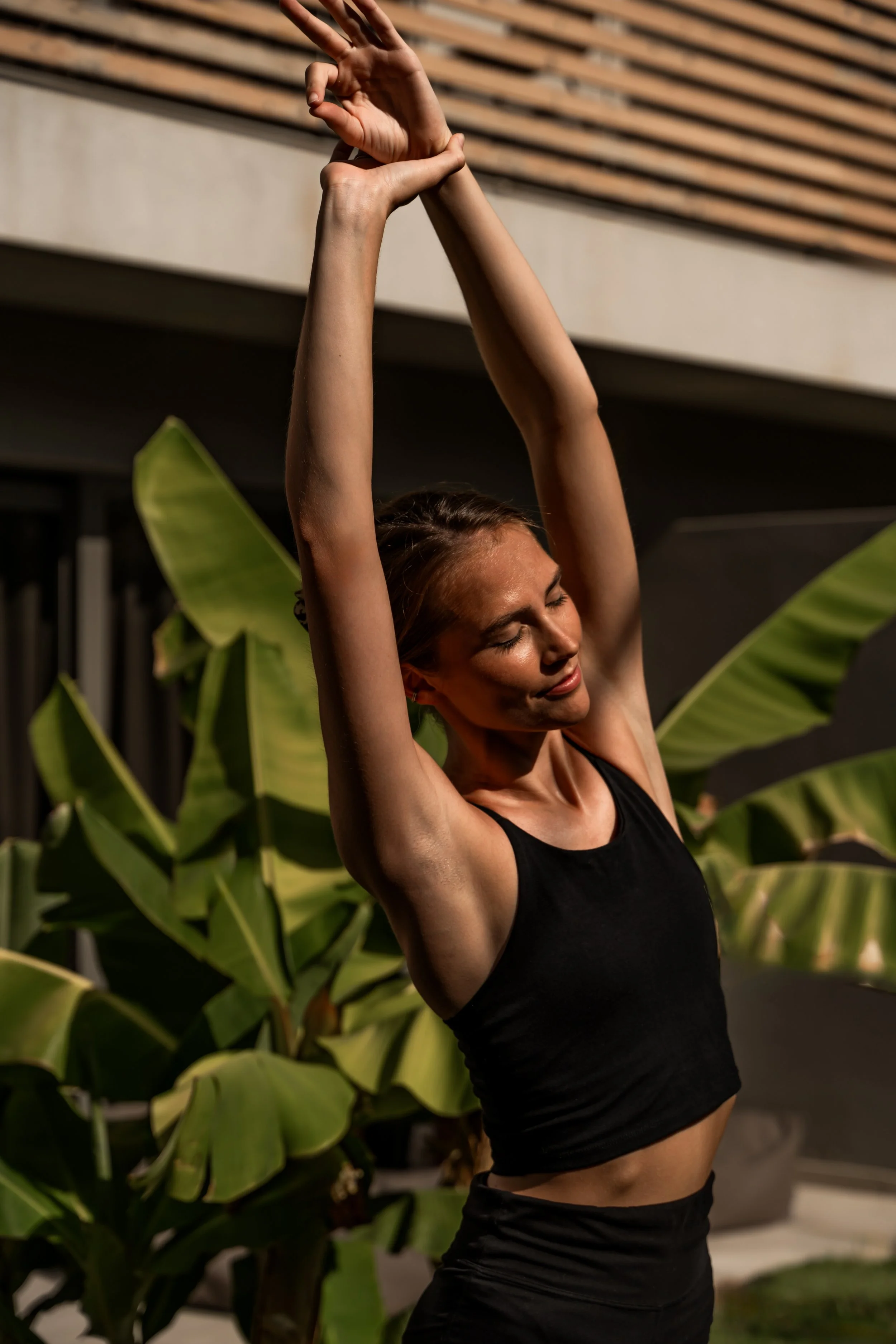 A woman in a black tank top and black workout shorts is stretching outdoors at night, with her arms raised above her head and eyes closed, standing in front of large green tropical plants.
