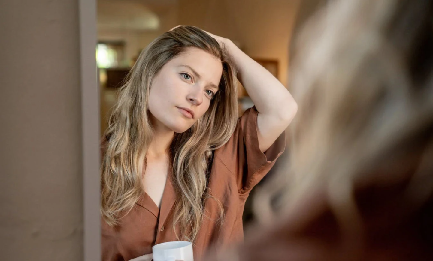 A woman with long wavy blonde hair, wearing a brown shirt, sitting indoors, looking at herself in a mirror, holding a white cup in her hand.