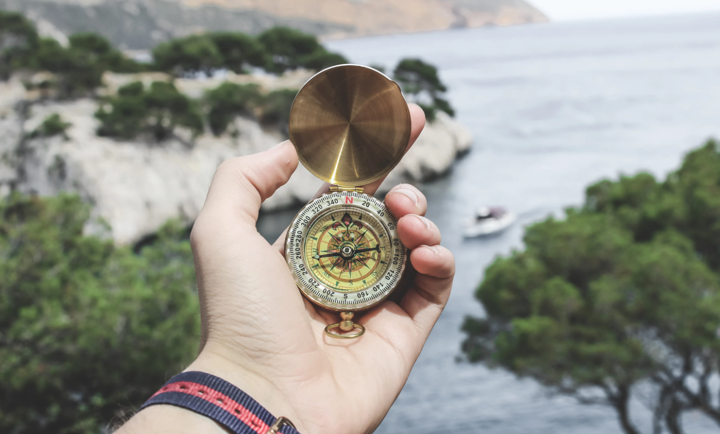 A person's hand holding a brass compass with open lid, pointing towards a scenic landscape of water, rocky cliffs, and green trees, with a boat on the water in the background.