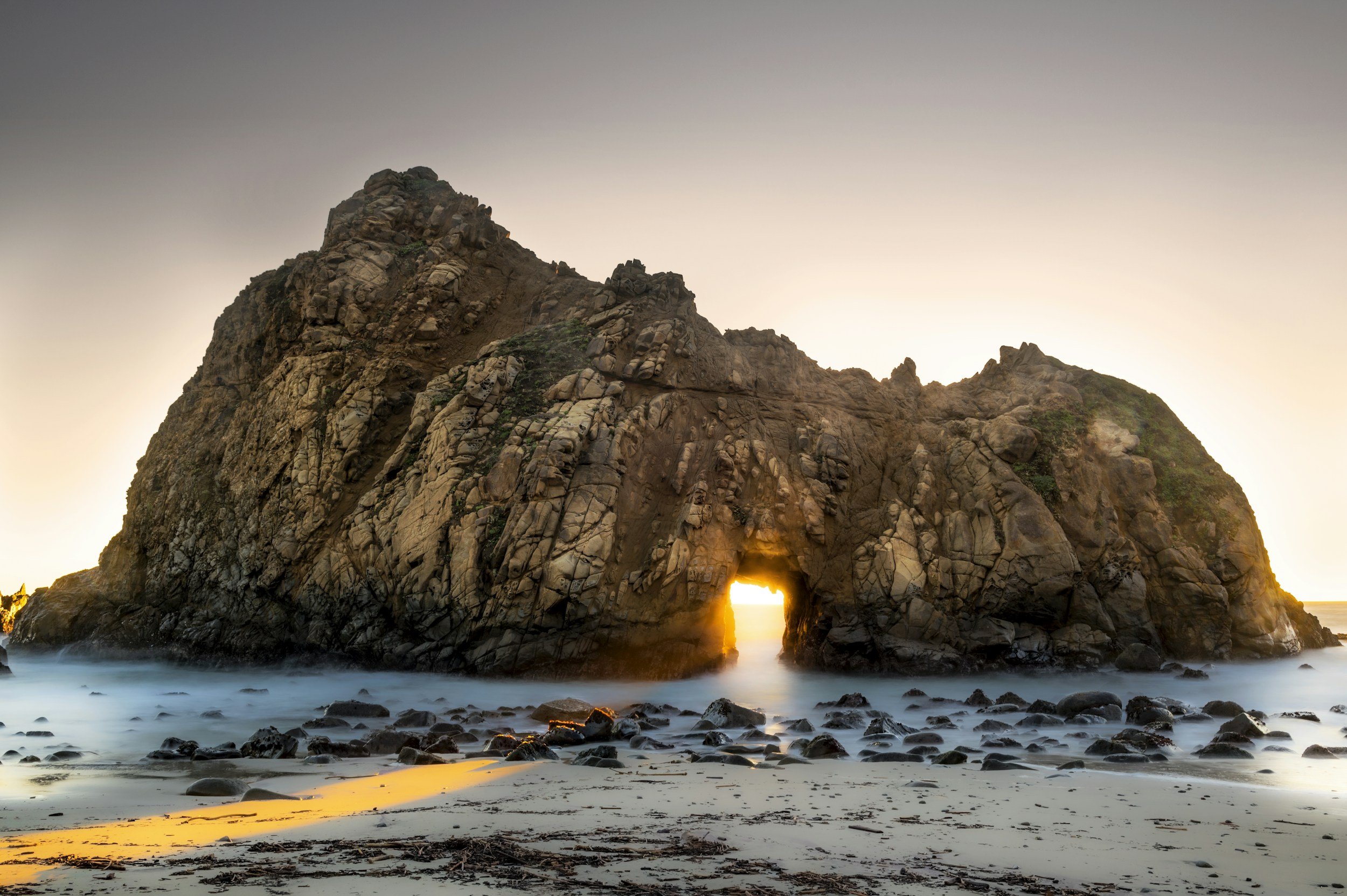 Rock formation with an arch at sunset, overlooking the ocean with a sandy beach and rocks in the foreground.