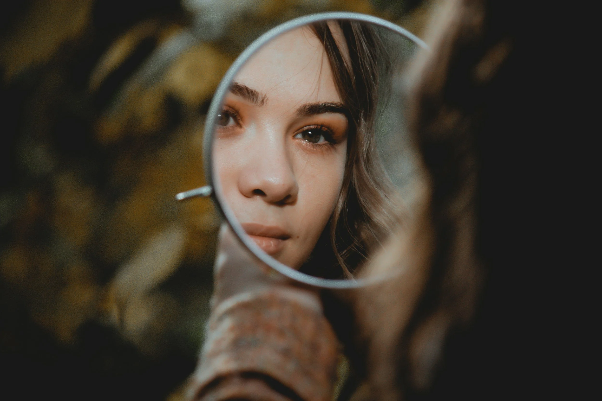 Close-up of a woman's face reflected in a small handheld mirror, with part of her long hair and shoulder visible. The focus is on her eyes and eyebrows.