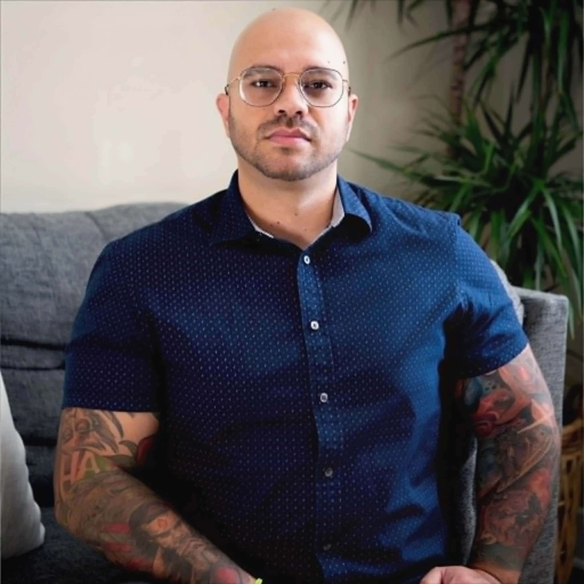 A man with glasses and a shaved head wearing a blue polka-dot shirt, sitting on a gray couch, displaying colorful tattoos on his arms, in a room with a large green garden plant in the background.