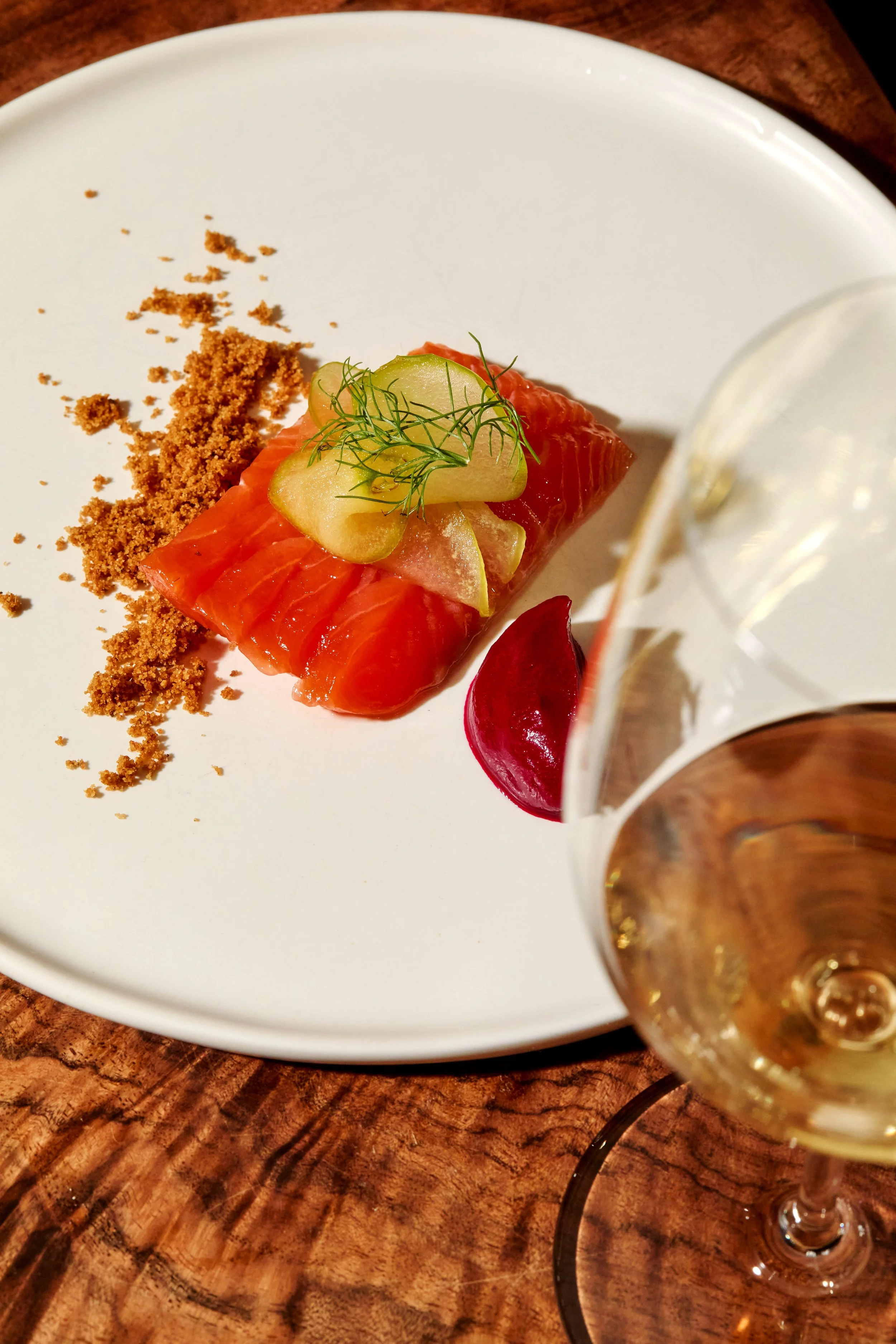 A plate with sliced salmon, topped with cucumber and dill, served with a beet slice and a pile of brown crumbs, with a glass of white wine in the foreground.