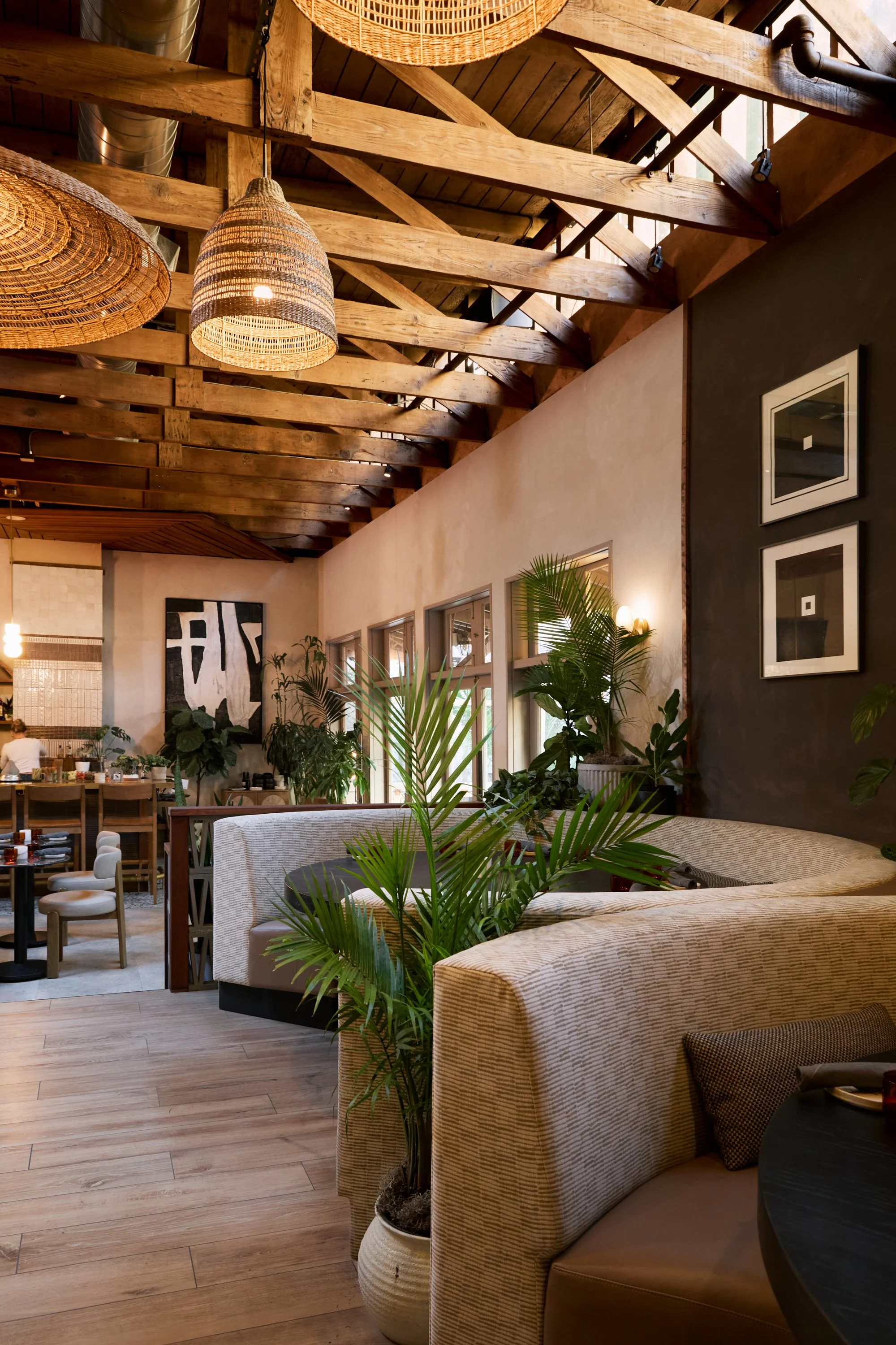 Interior of a cozy restaurant or cafe with wooden beams on the ceiling, hanging woven light fixtures, and lush green plants near windows, featuring beige upholstered seating and modern artwork on the walls.